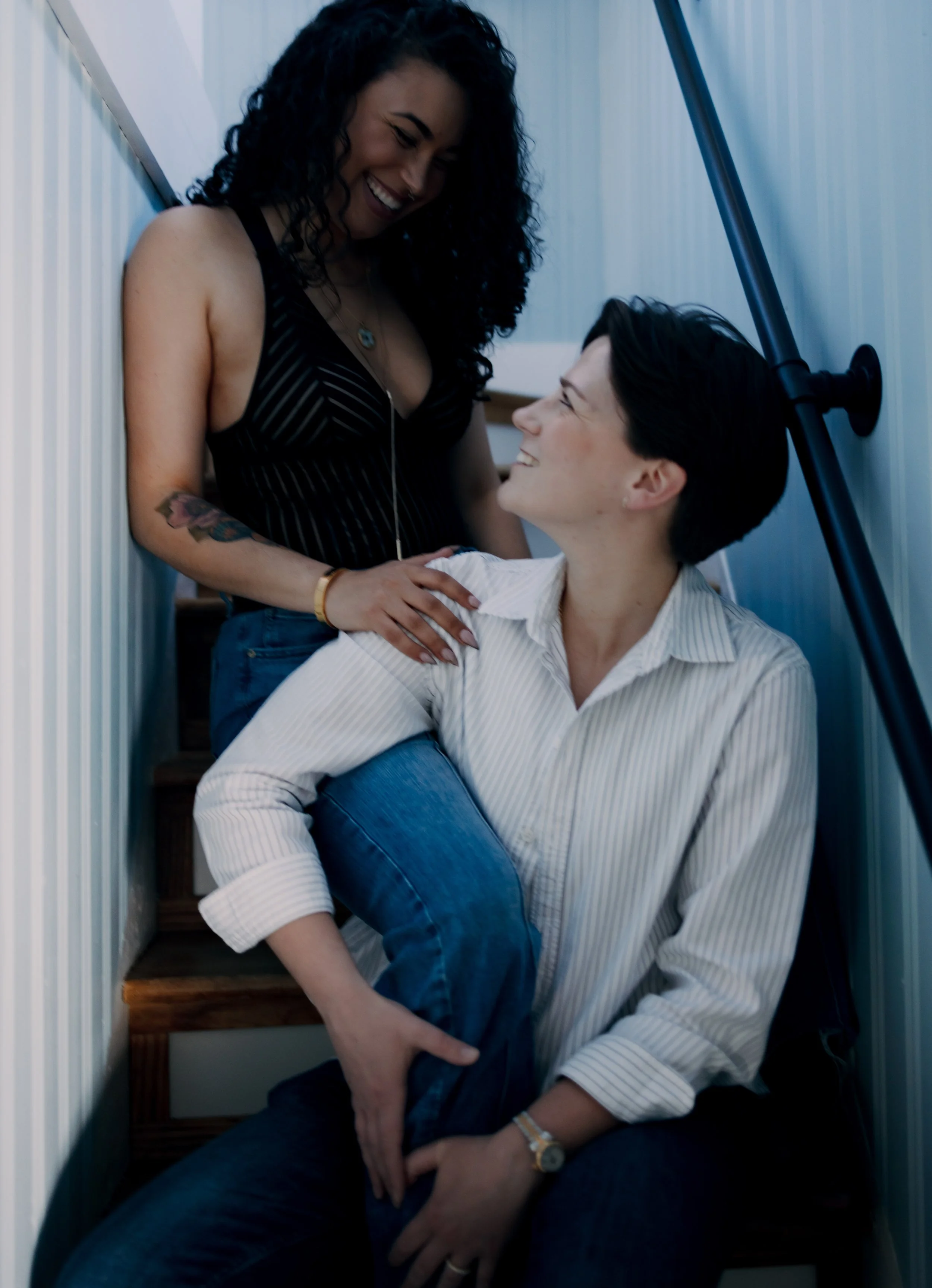 Two women sharing a joyful moment on a staircase, one sitting with her leg over the other's lap, both looking at each other and smiling.