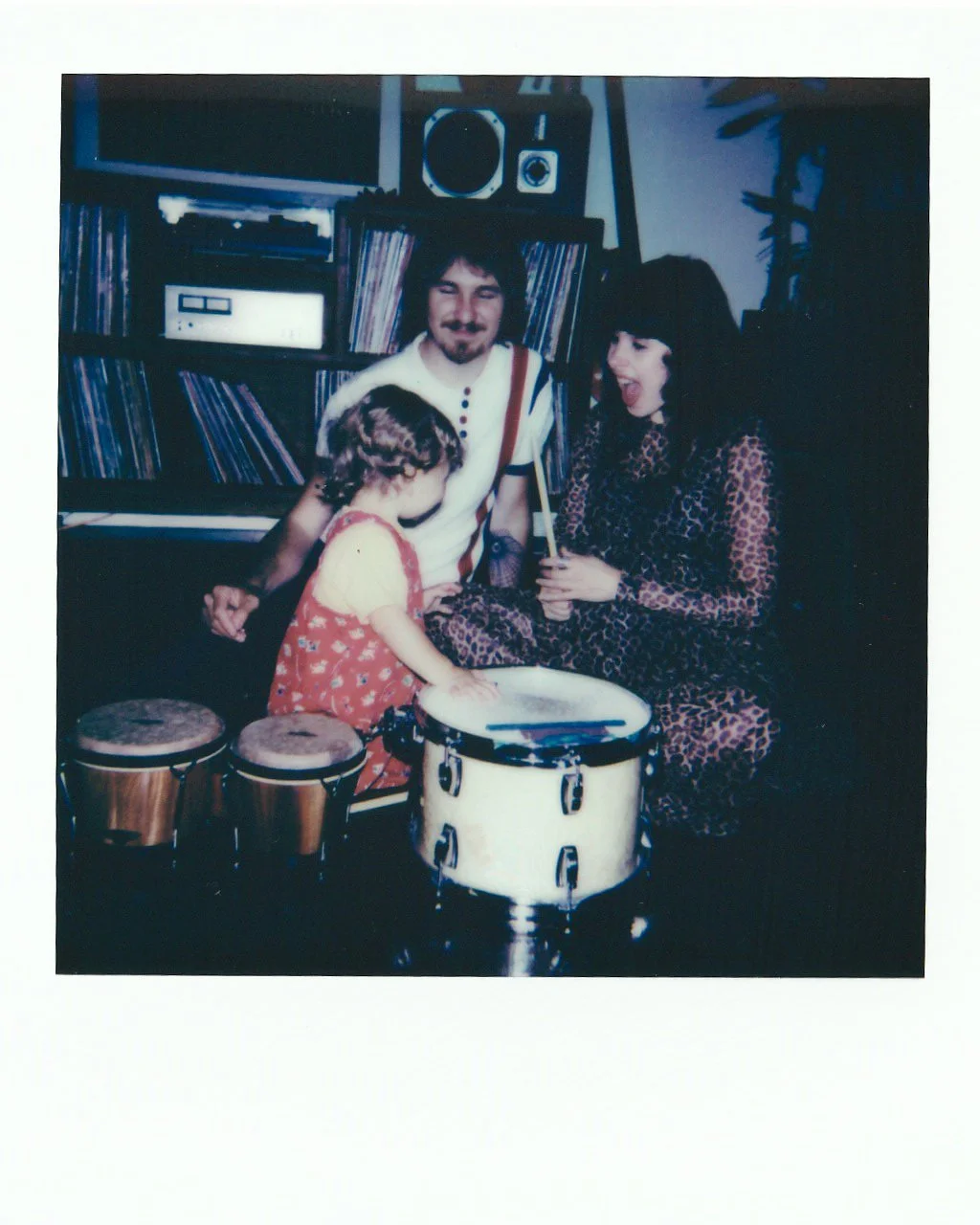 Three people, a man, a woman, and a young girl, are gathered around a drum set, smiling and playing together in a room with shelves of records and stereo equipment. The man has medium-length hair and a mustache, the woman has long dark hair, and the 