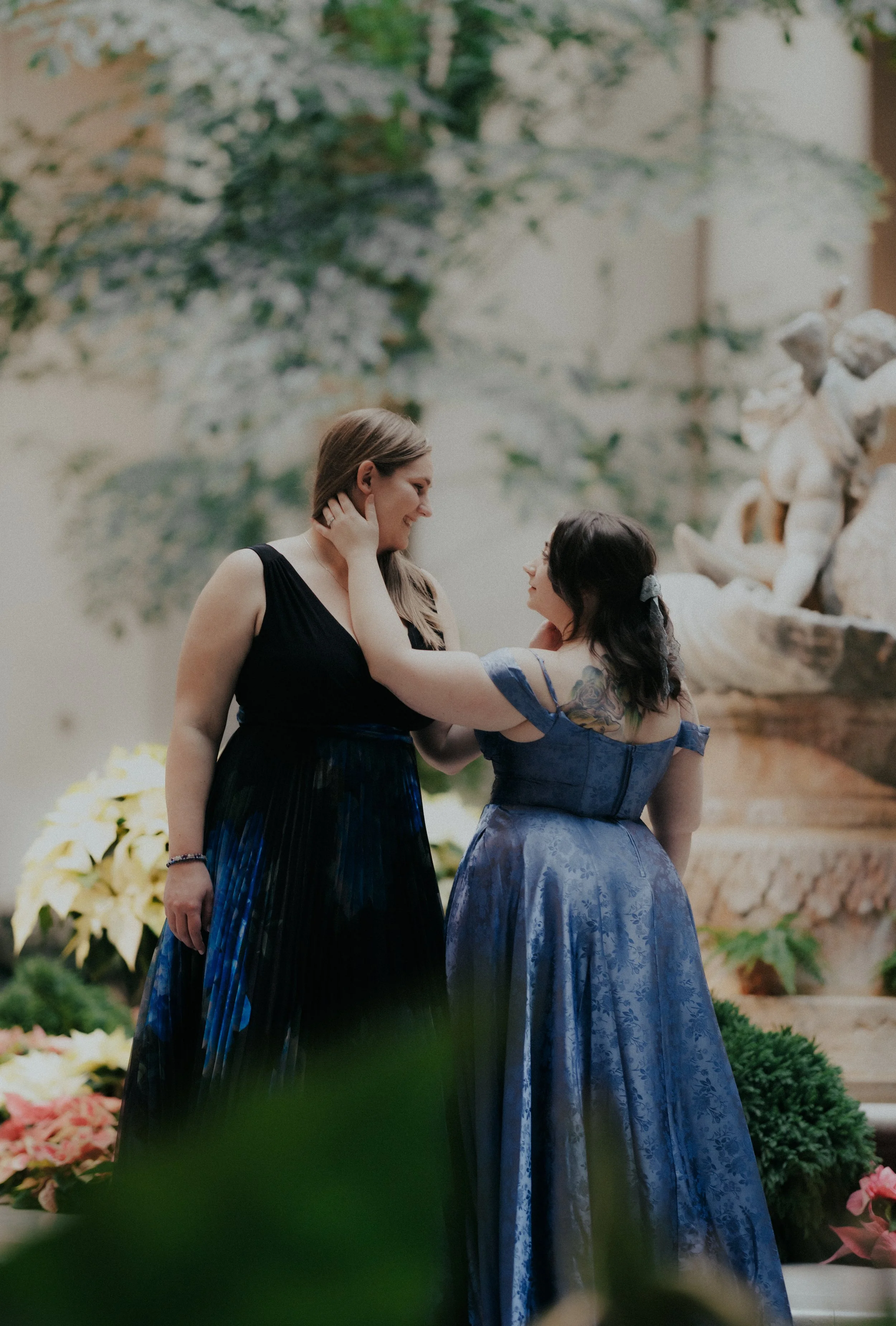 Two women in elegant dresses share a tender moment indoors, with flowers and a decorative fountain in the background.