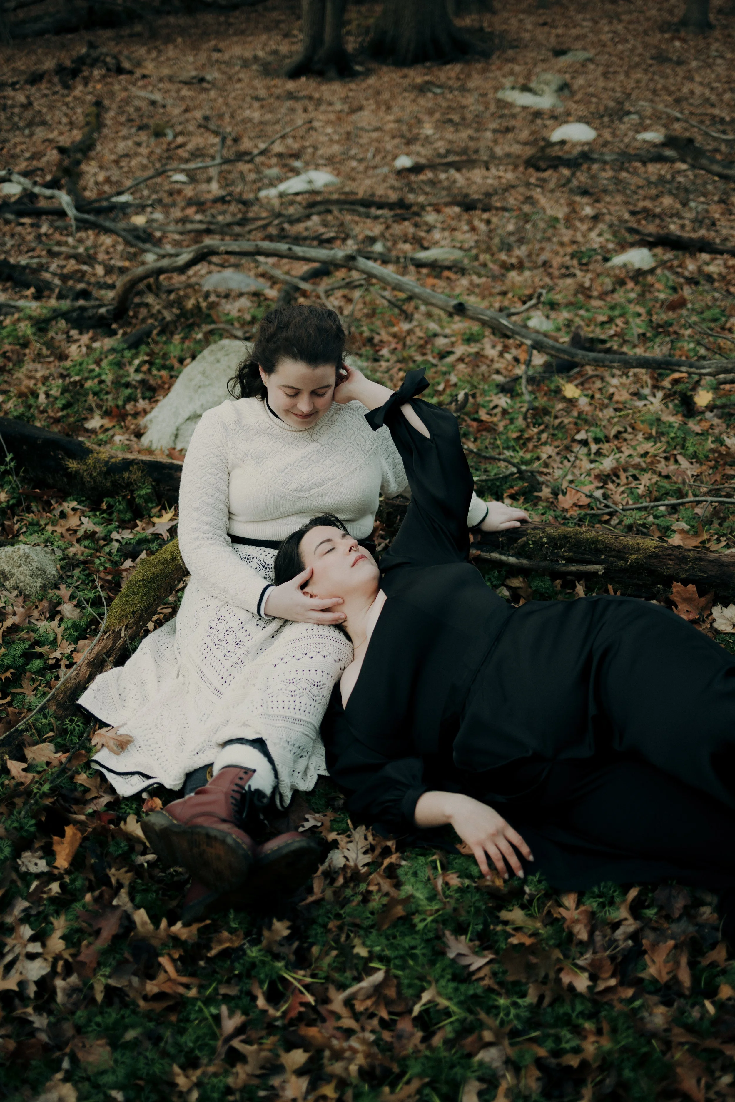 Two women lying on the forest floor among fallen leaves and branches, one resting her head on the other's lap, both appearing peaceful and relaxed.