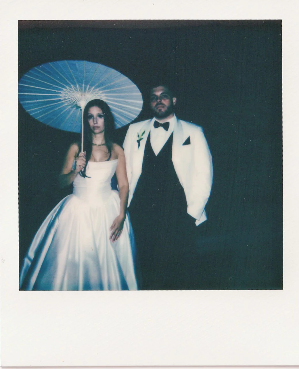 A man and woman dressed in formal attire, standing against a black background. The woman is wearing a white strapless gown and holding a blue parasol, with long dark hair. The man is wearing a white tuxedo jacket with black lapels, a black bow tie, and black pants, with a boutonniere on his lapel and a beard.