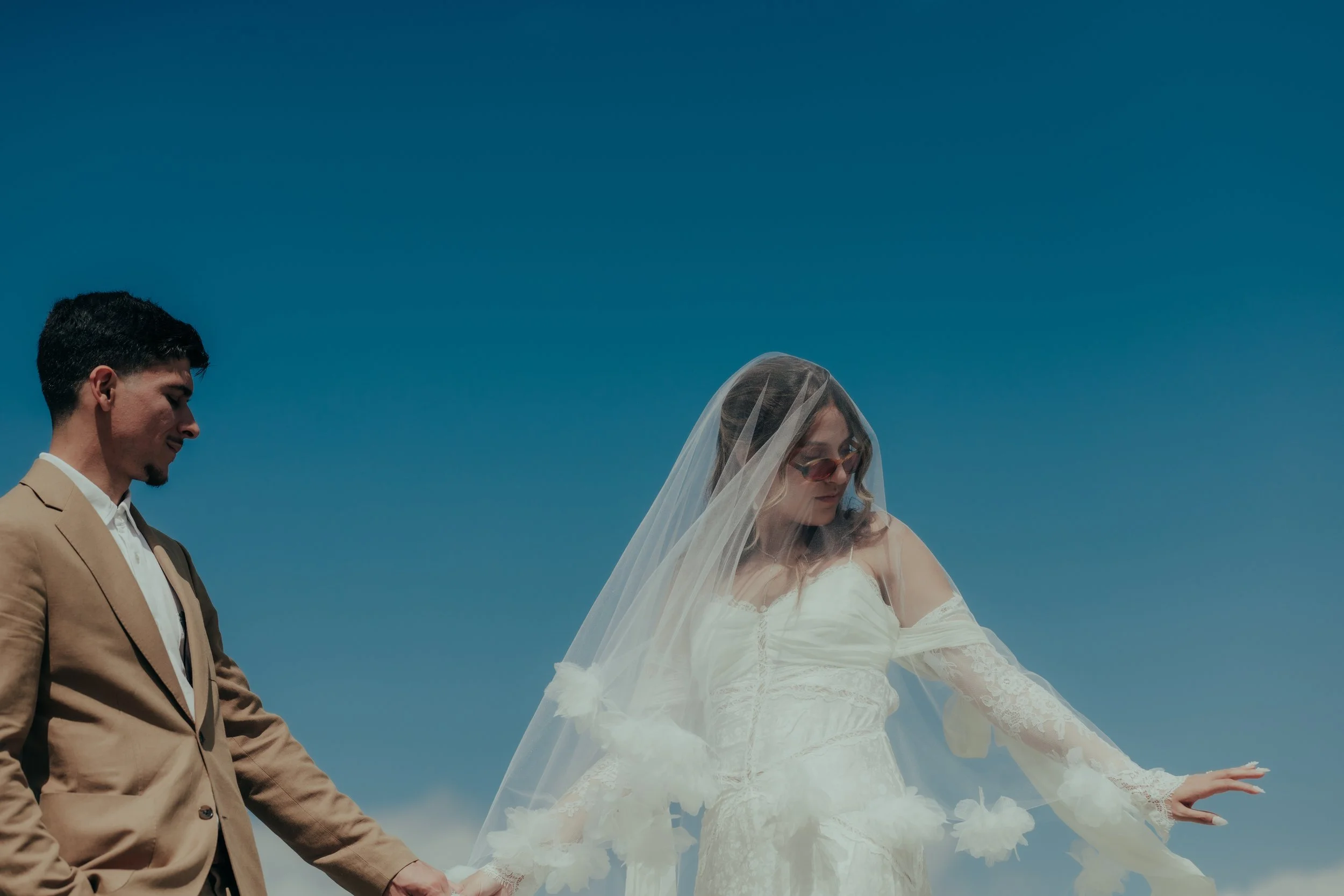 A newlywed couple holding hands outdoors against a bright blue sky, with the bride in a white wedding dress and veil, and the groom in a beige suit.