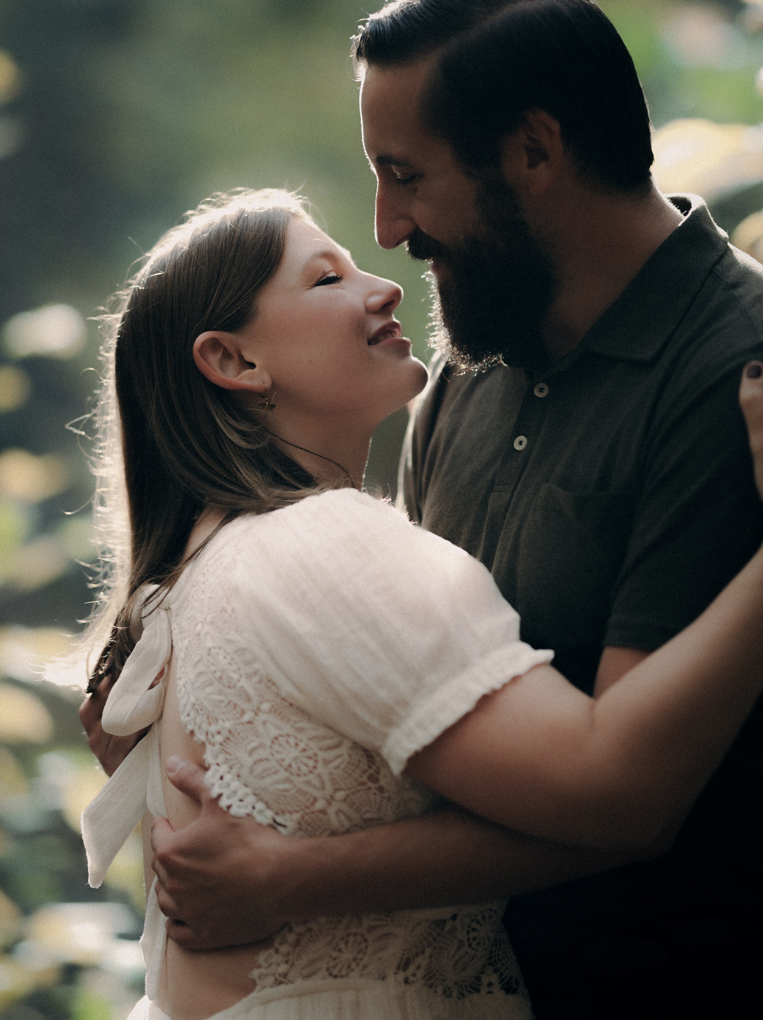 A man and woman are embracing and touching foreheads outdoors with sunlight filtering through trees in the background.