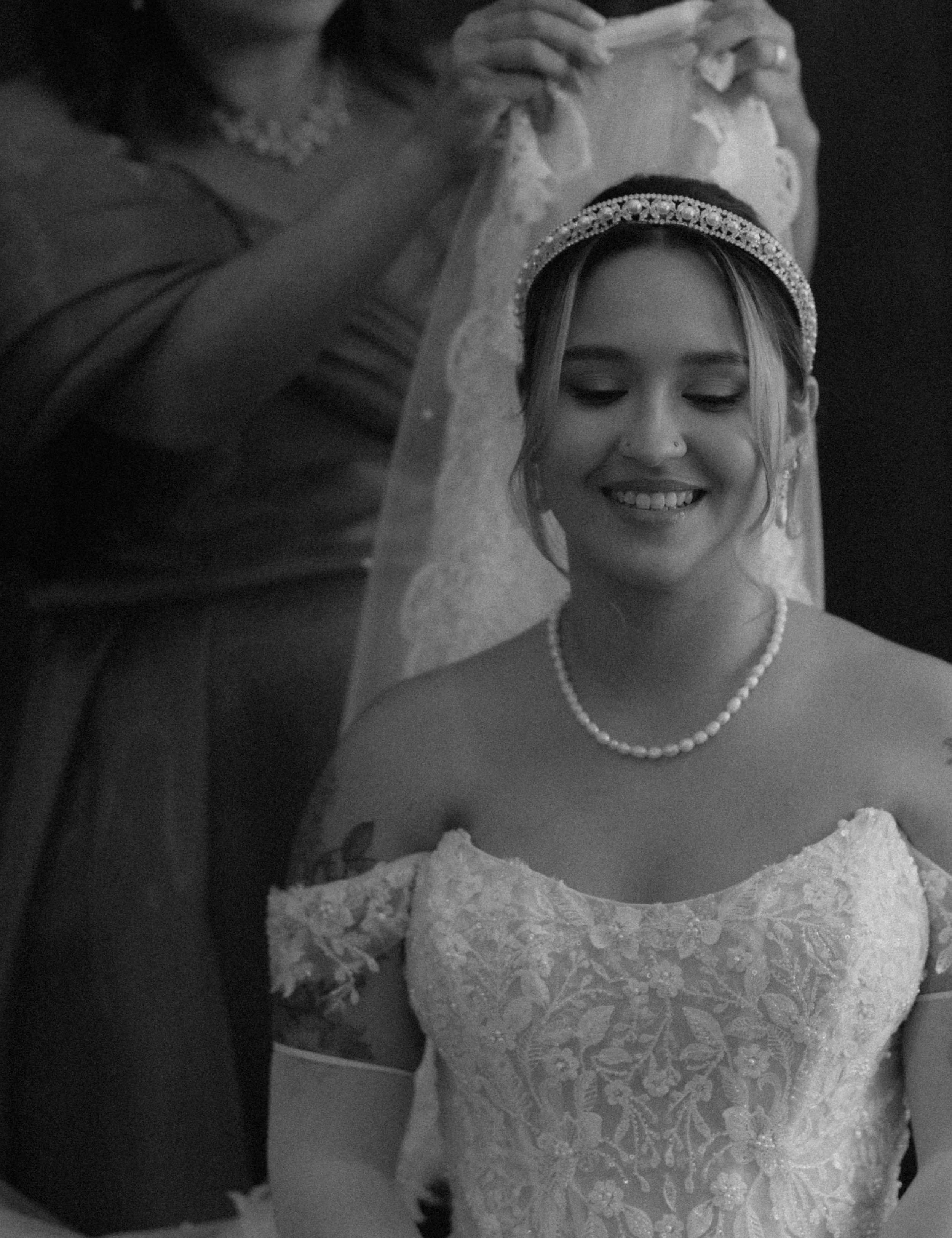 A bride smiling with her eyes closed, wearing a lace wedding dress, pearl necklace, and a bejeweled headband. An older woman adjusts her veil in the background.