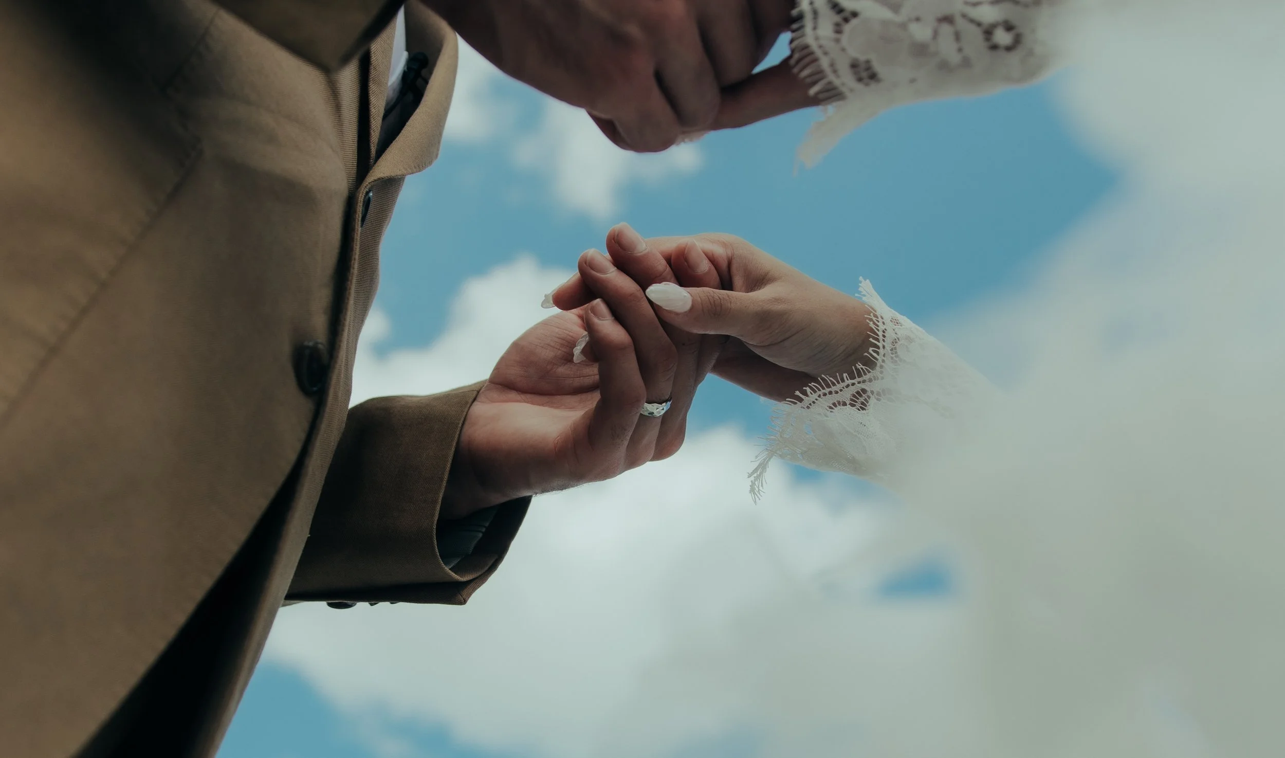 A couple holding hands in a wedding ceremony, with the sky and clouds in the background.