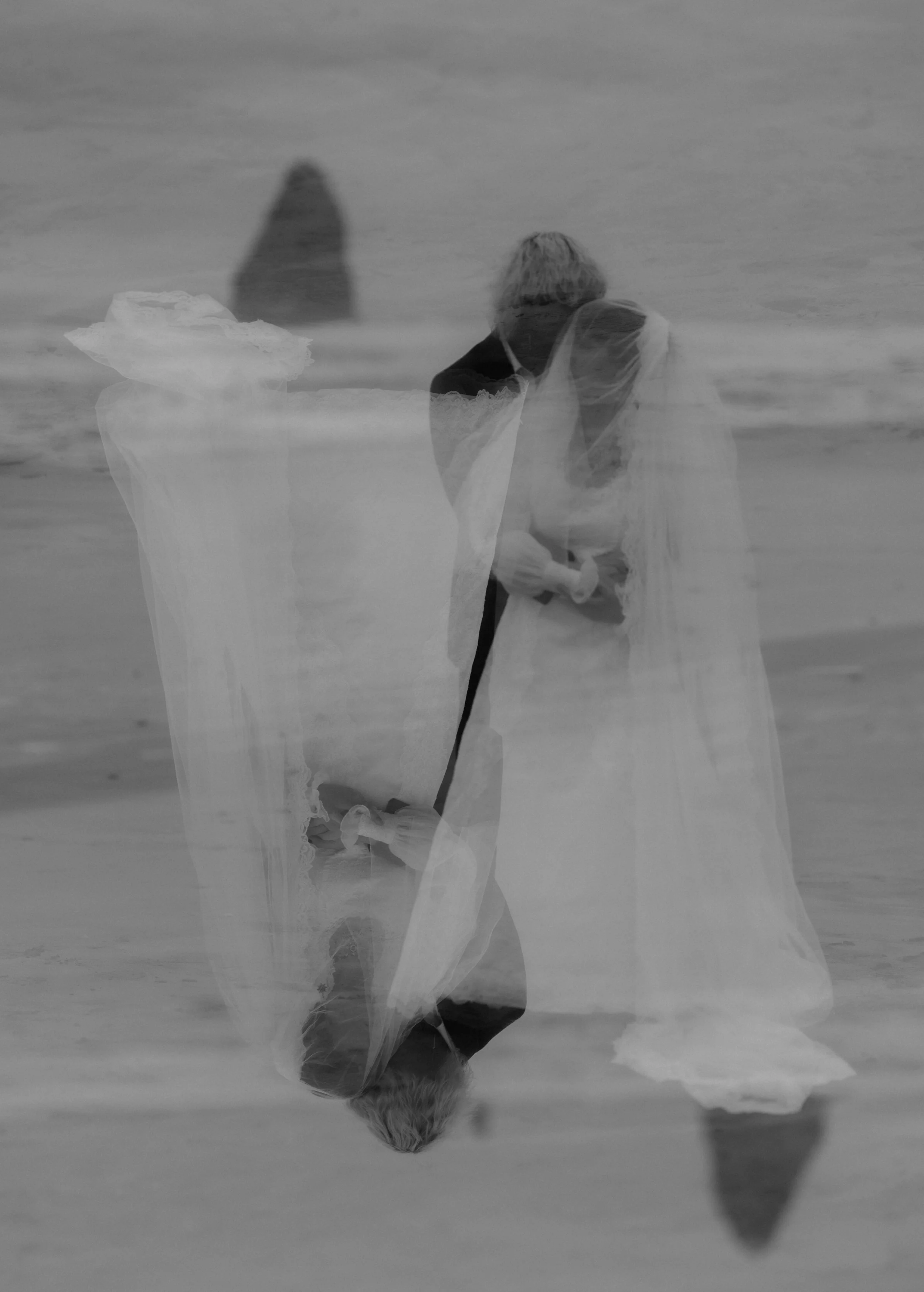A black and white photo of a same-sex wedding ceremony on the beach with two women in wedding dresses and an officiant, reflected in the wet sand.