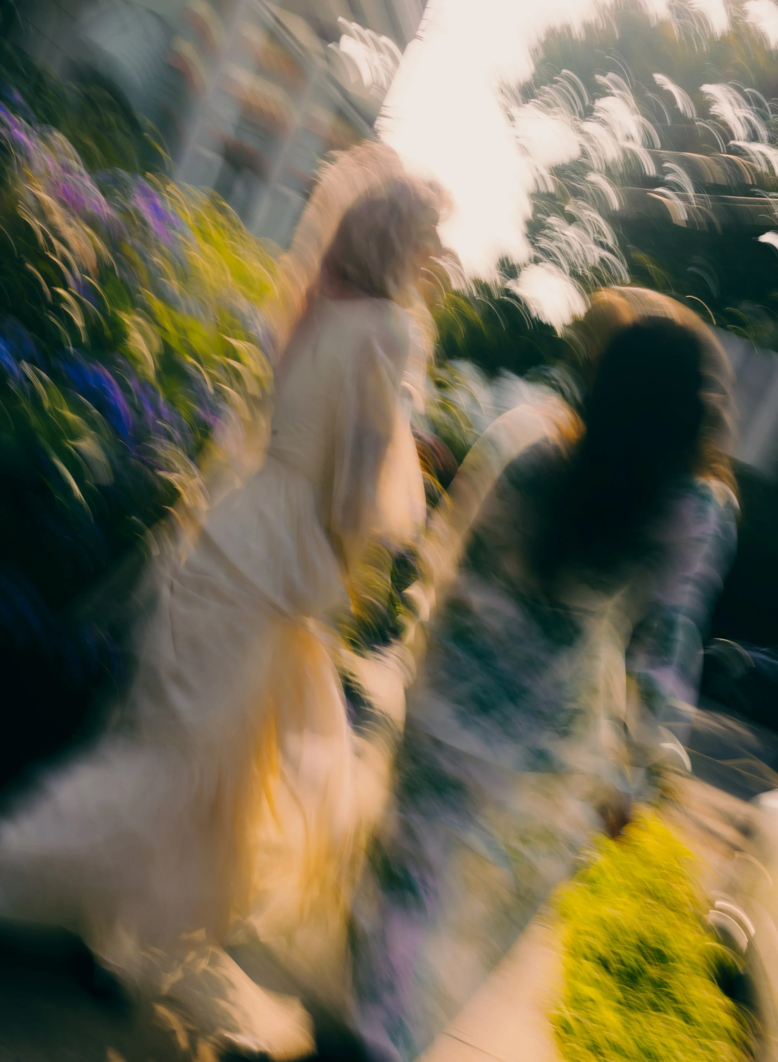Blurred photo of two women, one standing and one sitting, outdoors with flowers and greenery, during sunset.