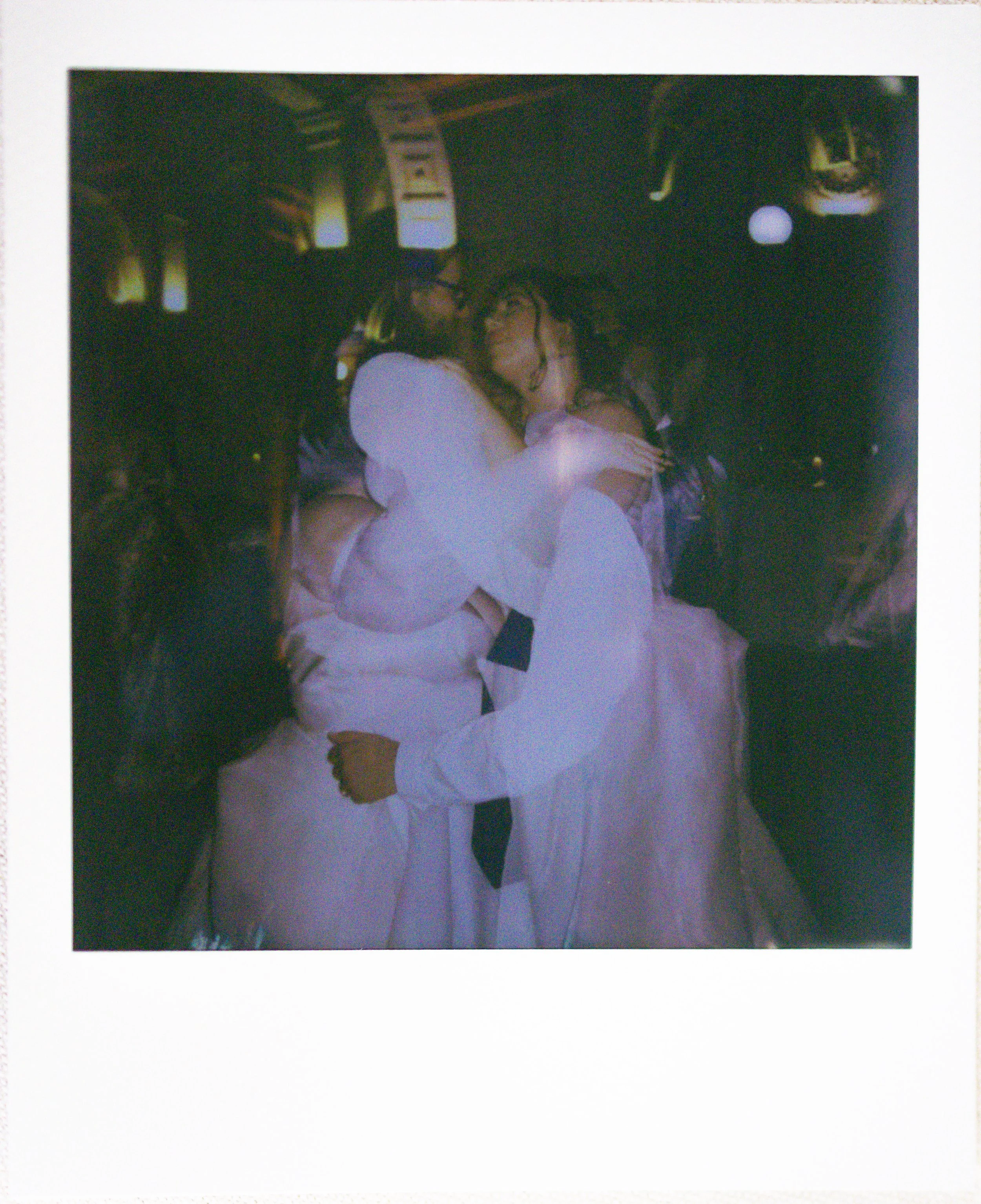 Two women in white wedding dresses sharing a kiss on the dance floor.