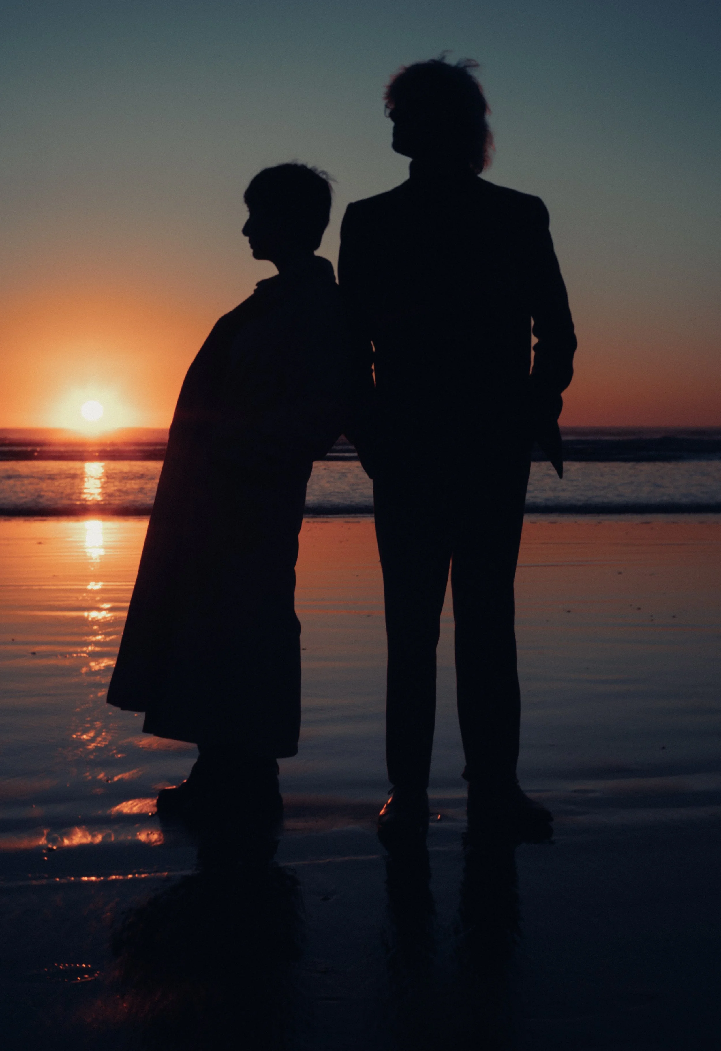Silhouettes of two people, a woman and a girl, standing on the beach during sunset.