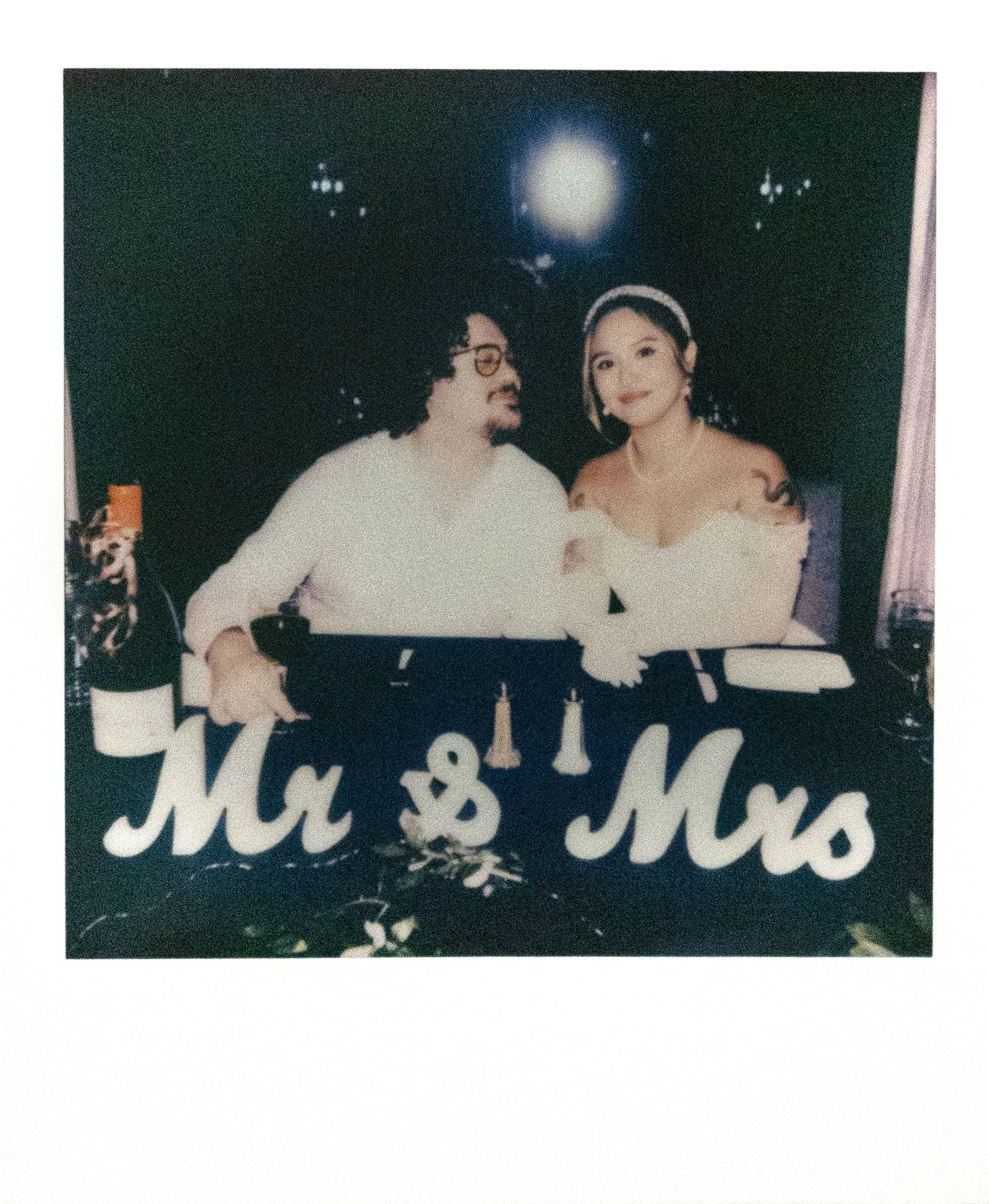 A couple sitting in front of a black backdrop with a moonlit scene, with a sign in front reading 'Ma & Mso', beer bottles on the table, and a woman in a white off-shoulder dress and headband posing with a man in glasses and a white shirt.