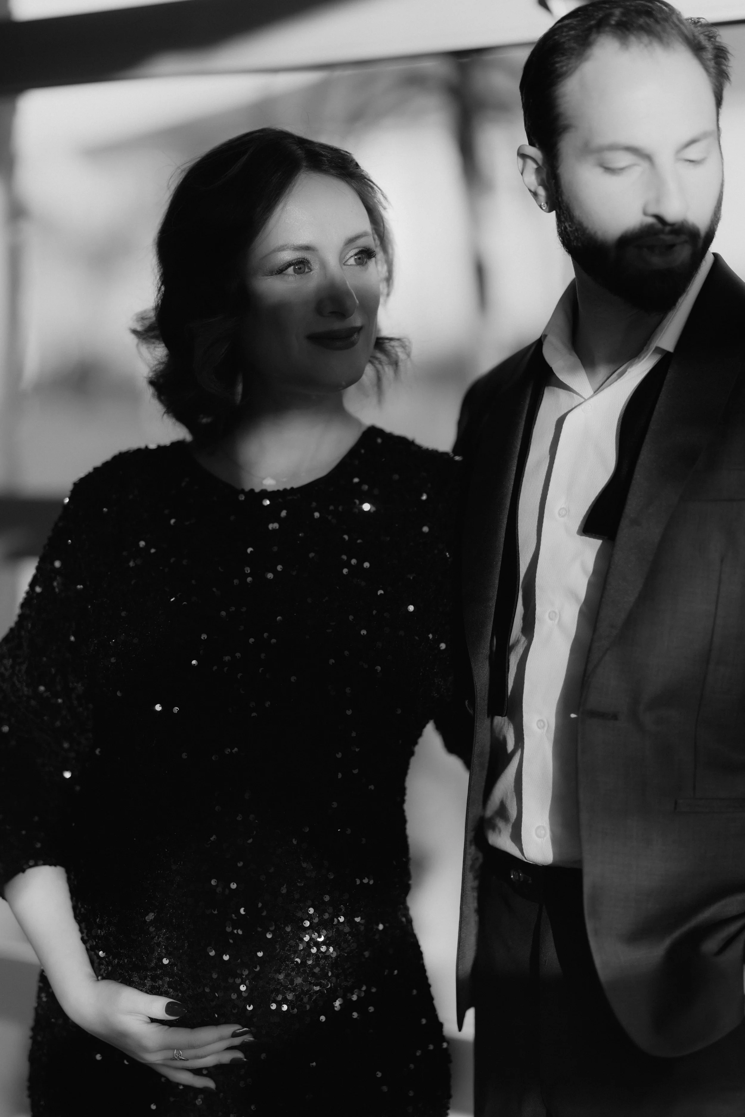 Black and white photo of a woman in a sequined dress and a man in a suit standing close together, with the woman gently holding her stomach and the man with eyes closed.