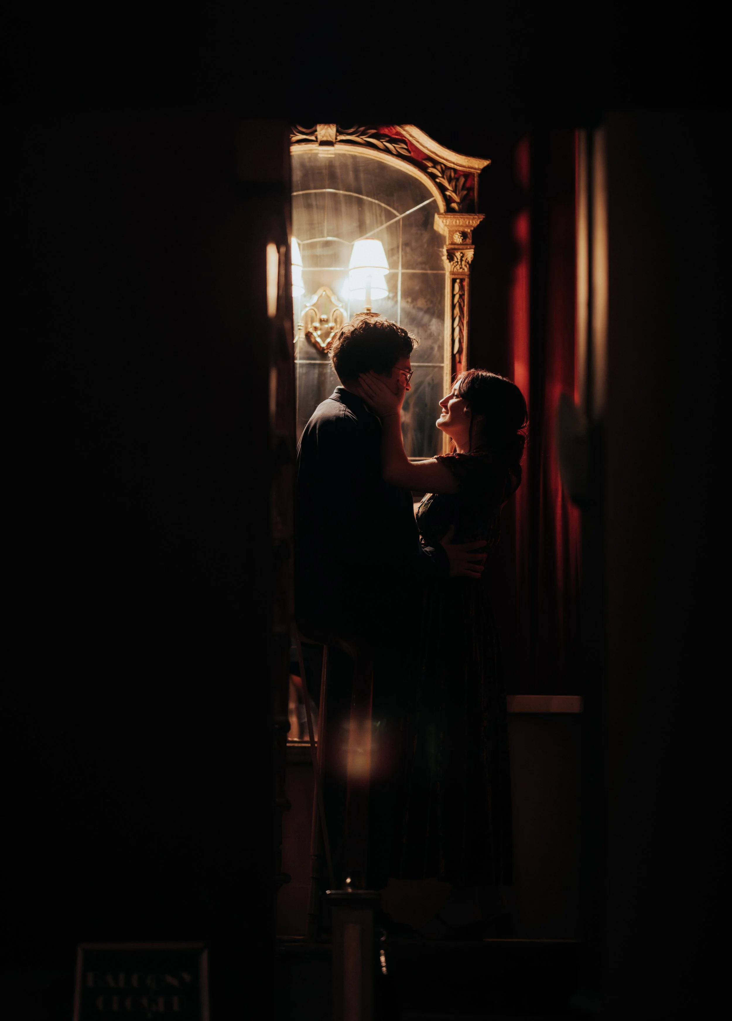 A couple sharing a romantic moment in dim lighting, with the woman holding the man's face, standing in front of an ornate mirror with warm lamps reflected behind them.
