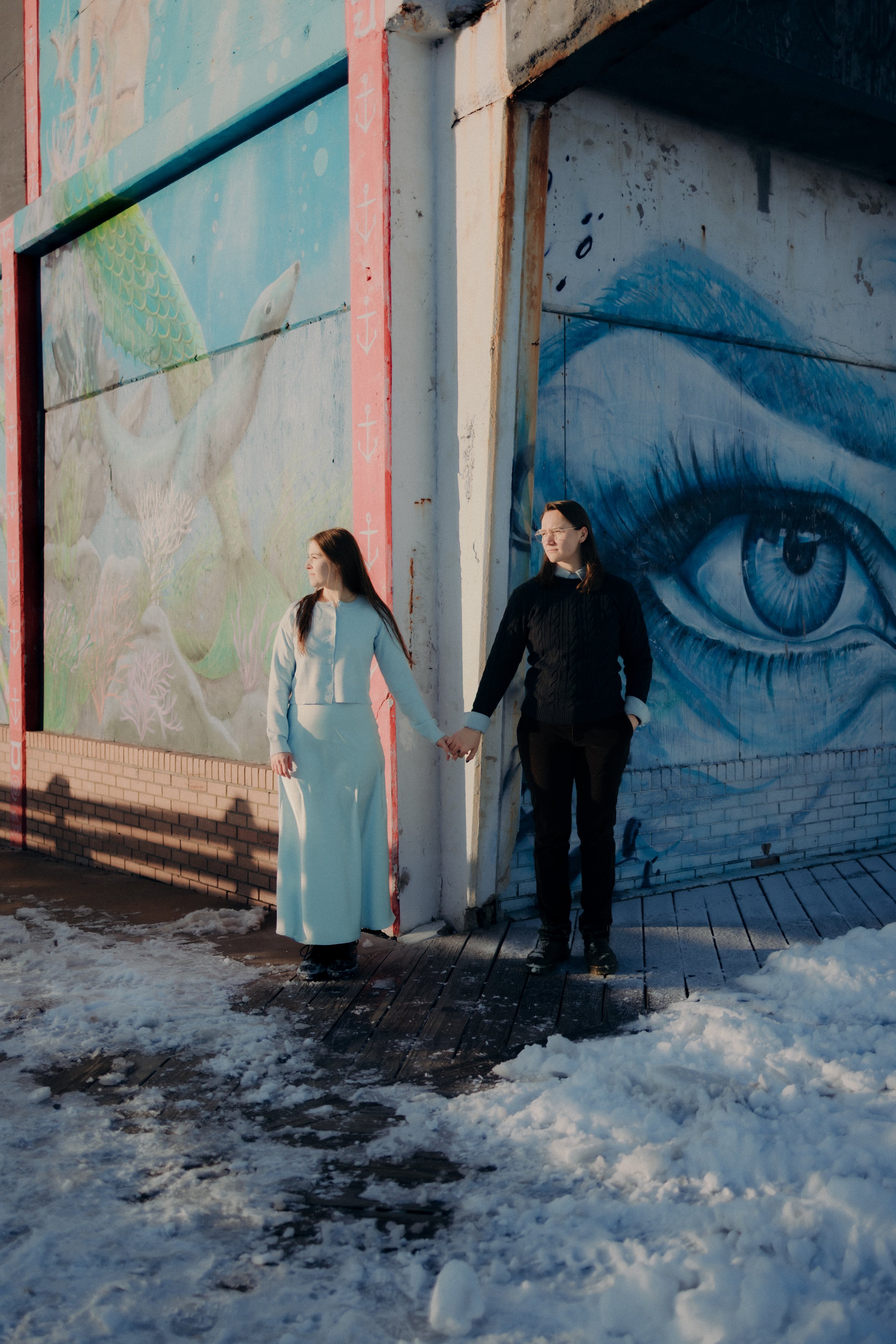 Two women standing hand in hand in front of a wall with colorful murals, one of a turtle and the other of an eye, on a snowy sidewalk during daytime.