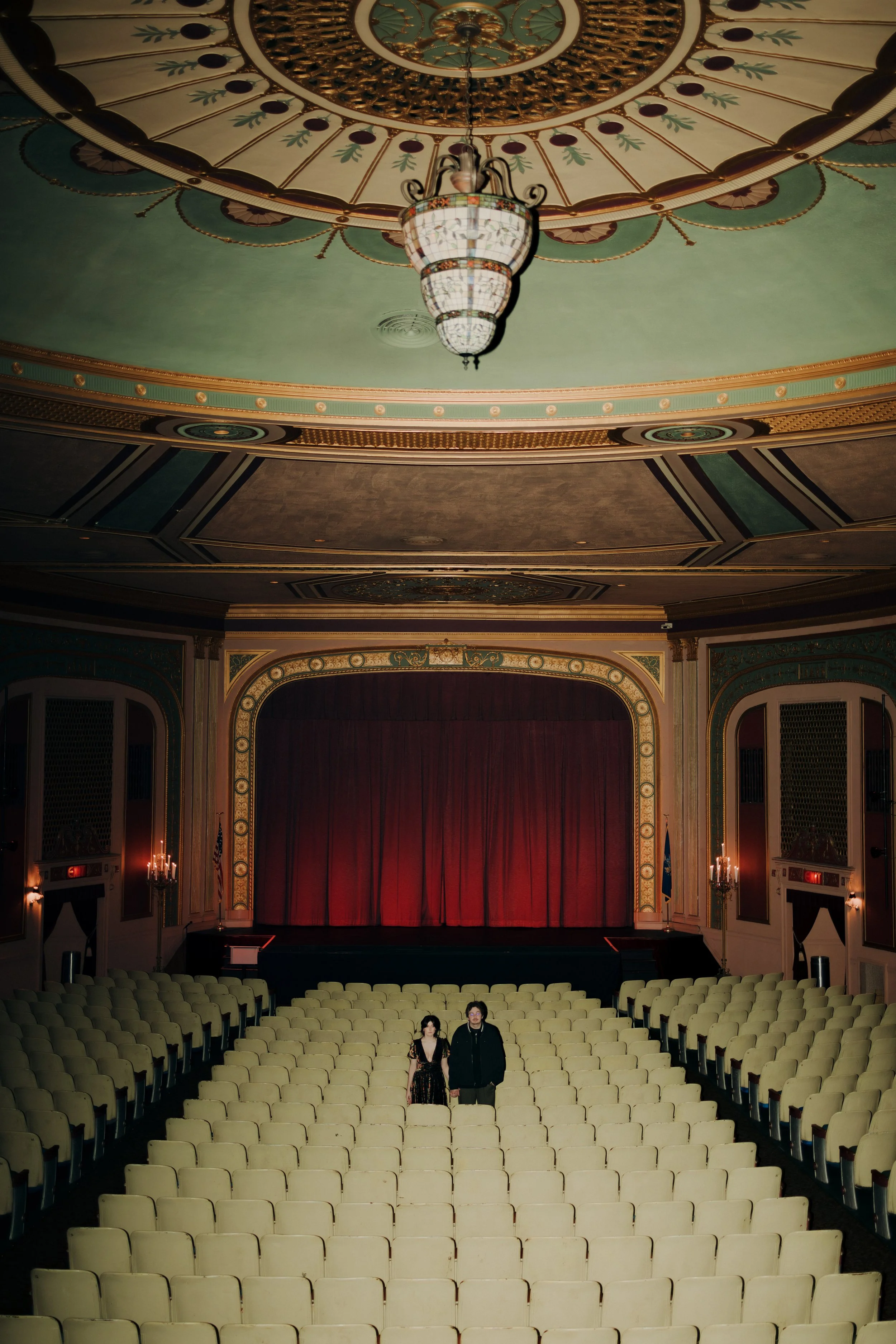 An ornate theater auditorium with rows of empty cream-colored seats, a stage with red curtains, and a couple standing in the center aisle.