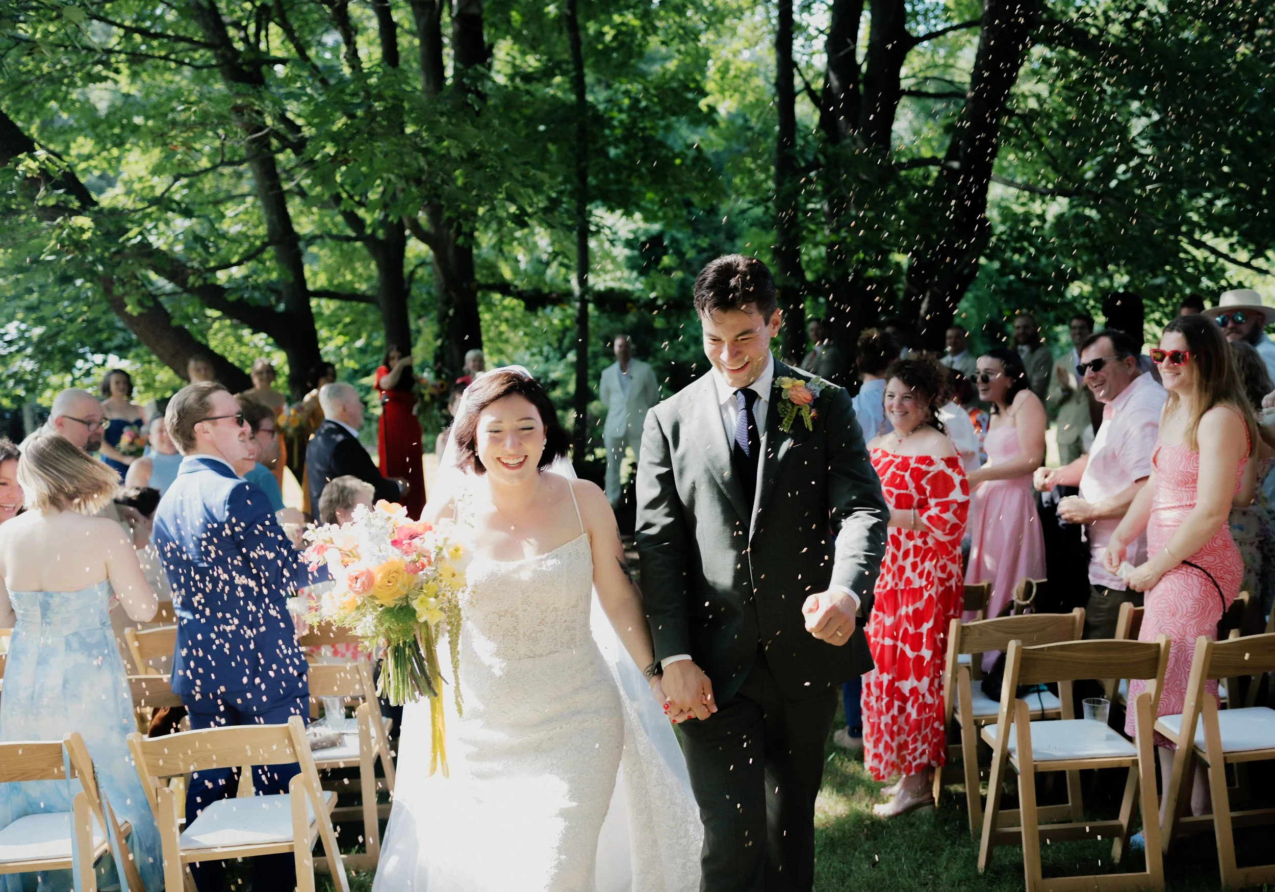 A newlywed couple walking hand in hand outdoors after their wedding ceremony, surrounded by friends and family who are celebrating and throwing confetti.