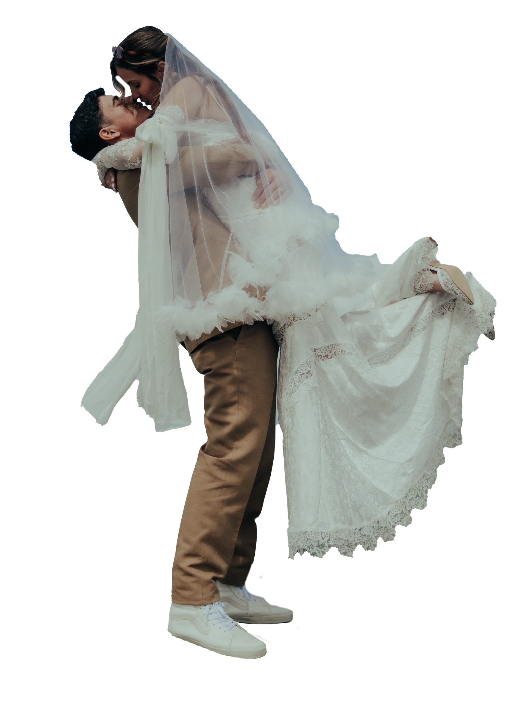 A groom in beige pants and white shoes lifting a bride in a wedding dress with lace details and a veil, holding her close in a romantic pose.