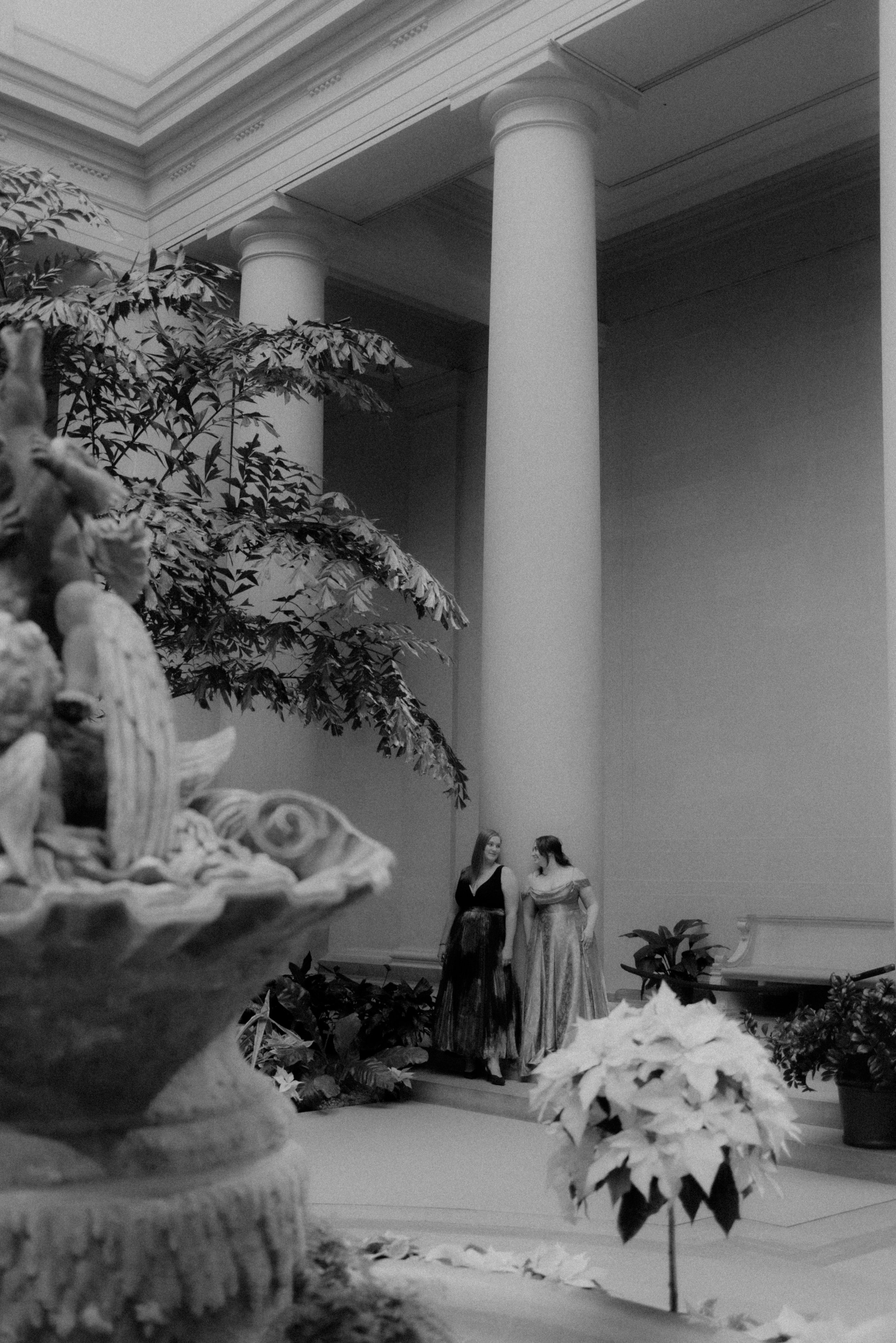 Two women in formal gowns standing and talking in front of a large room with tall columns and potted plants, with a decorative fountain in the foreground.