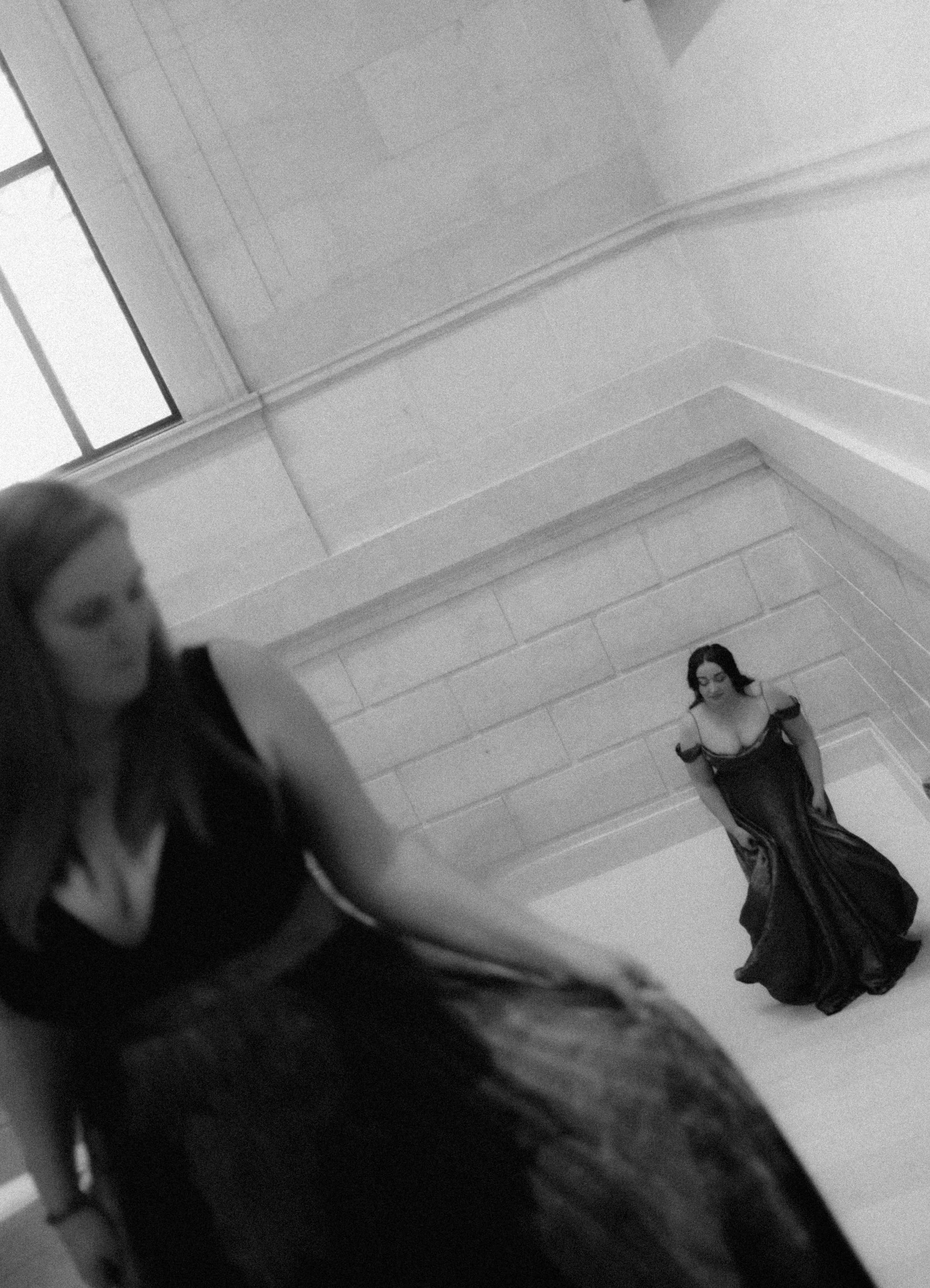 Two women in black dresses, one sitting on a ledge with an out-of-focus face in the foreground, the other standing against a wall, both in a modern, well-lit indoor space.