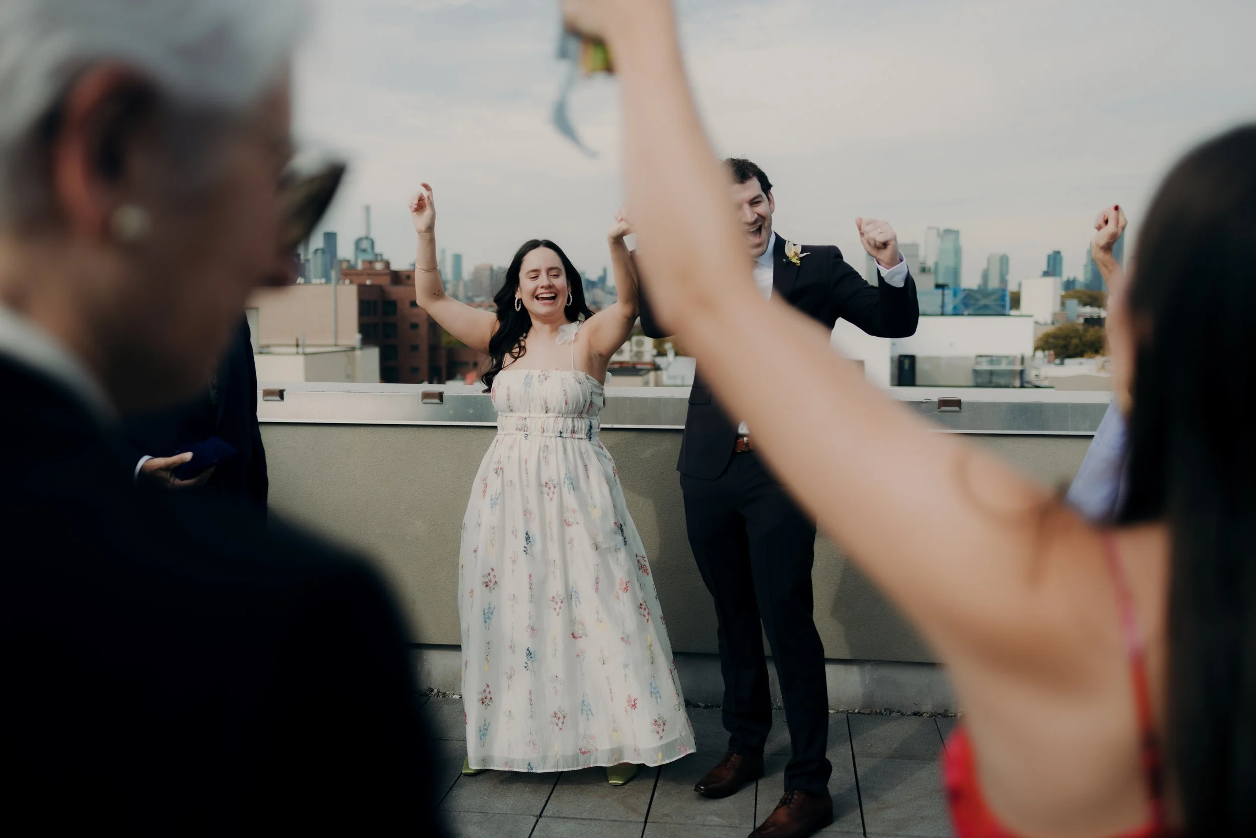 People celebrating on a rooftop with a city skyline in the background, smiling and raising their arms.