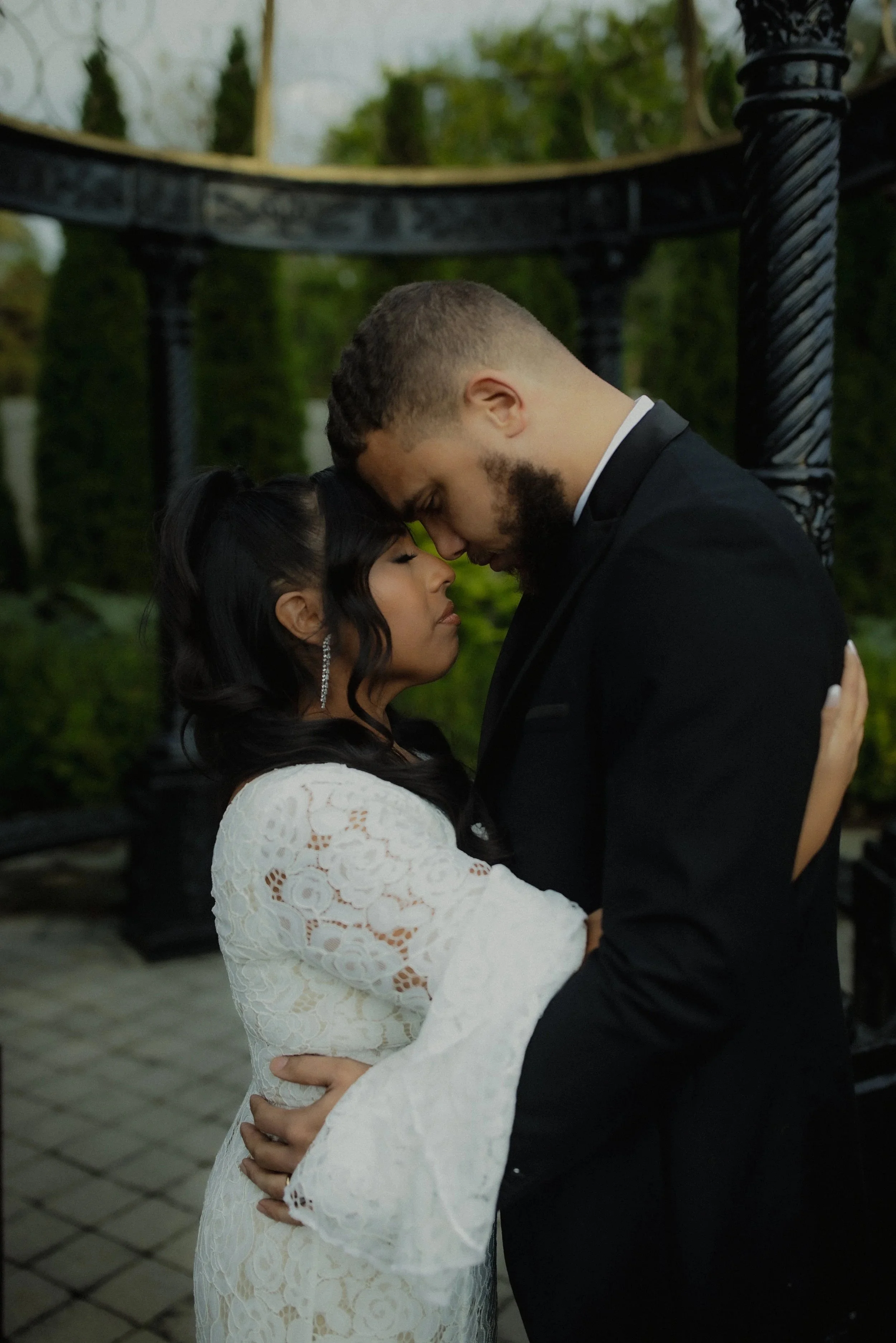 A couple dressed in wedding attire sharing an intimate moment, foreheads touching, outdoors with greenery and black metal furniture in the background.
