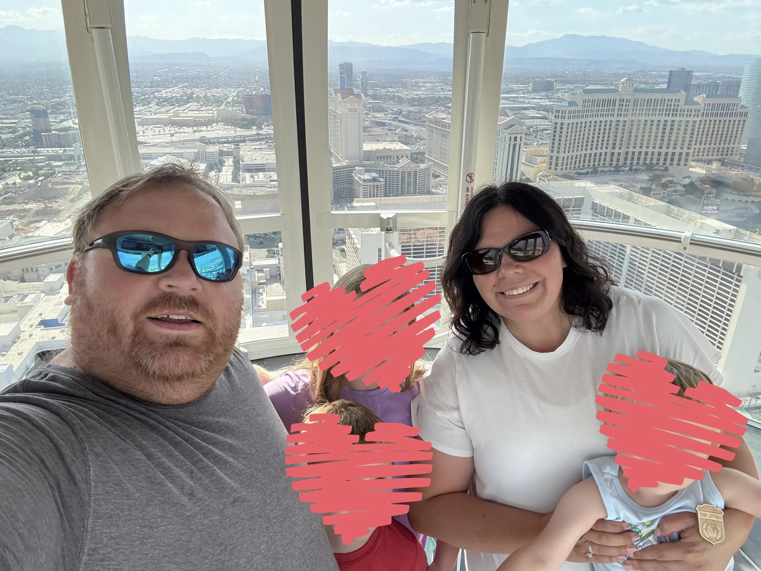 A family inside the high observation deck of the Eiffel Tower in Las Vegas, Nevada, with a cityscape view in the background. The man in the foreground is wearing sunglasses and a gray t-shirt, and the woman is wearing sunglasses and a white t-shirt, holding a young child. Two children with their faces covered are also present.