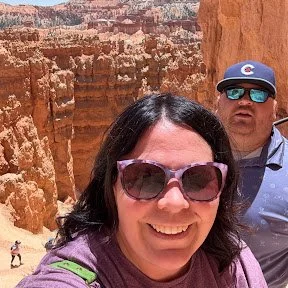 A woman smiling at the camera with sunglasses, outdoors at a canyon with orange rock formations, and a man wearing sunglasses and a cap standing in the background.