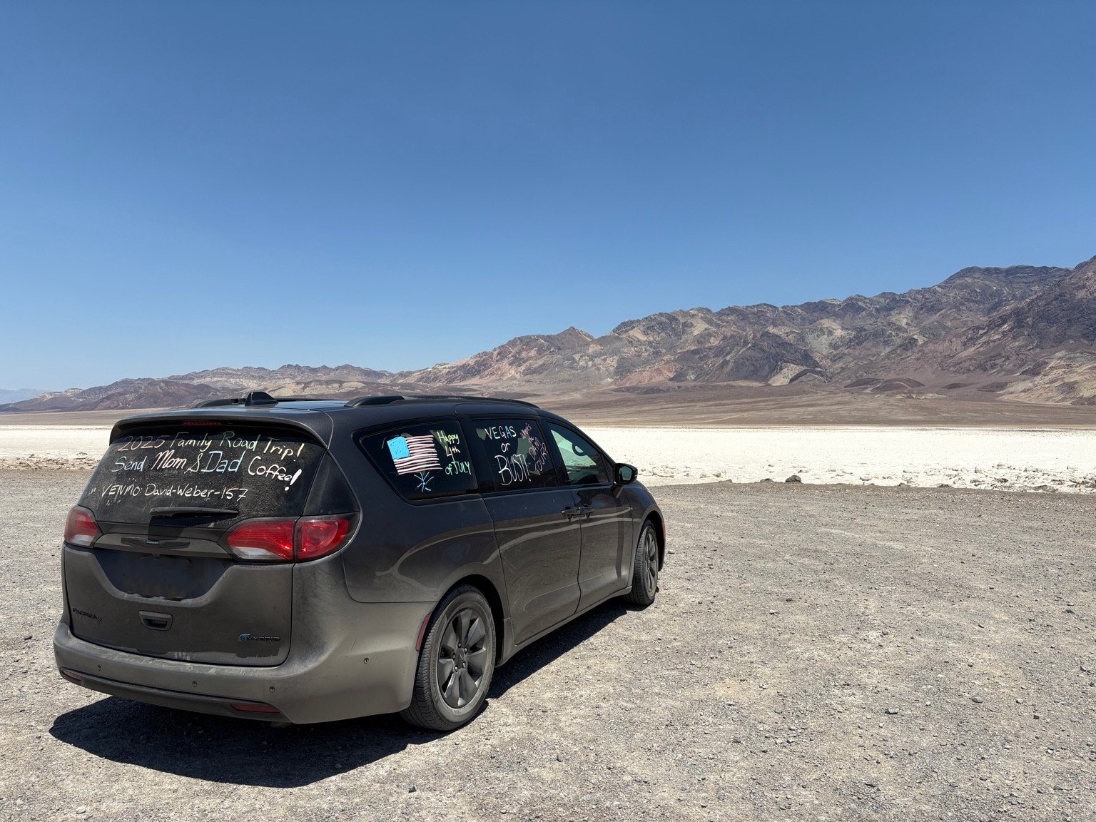 A black minivan parked on a desert landscape with mountains in the background. On the rear and side windows, messages and drawings are written in white and colored markers, including a U.S. flag, a cactus, and festive decorations.