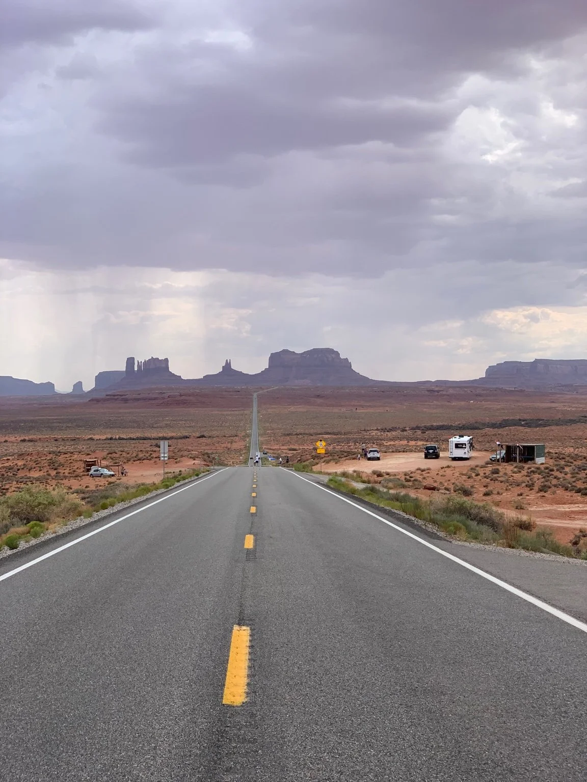 Long straight road in a desert landscape with mesas in the distance under a cloudy sky.