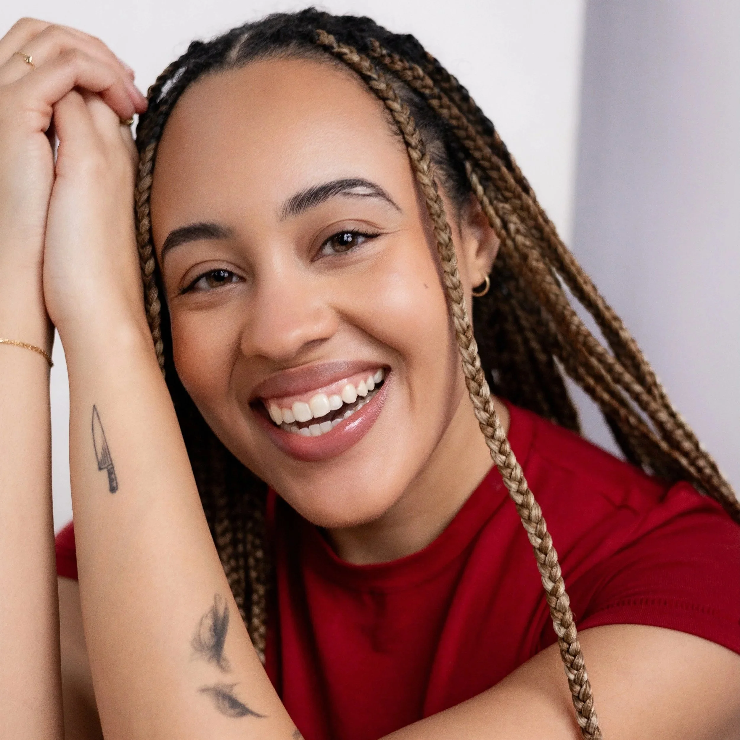 A smiling woman with braided hair, wearing a red shirt, showing tattoos on her arm, and posing with her hand near her head.