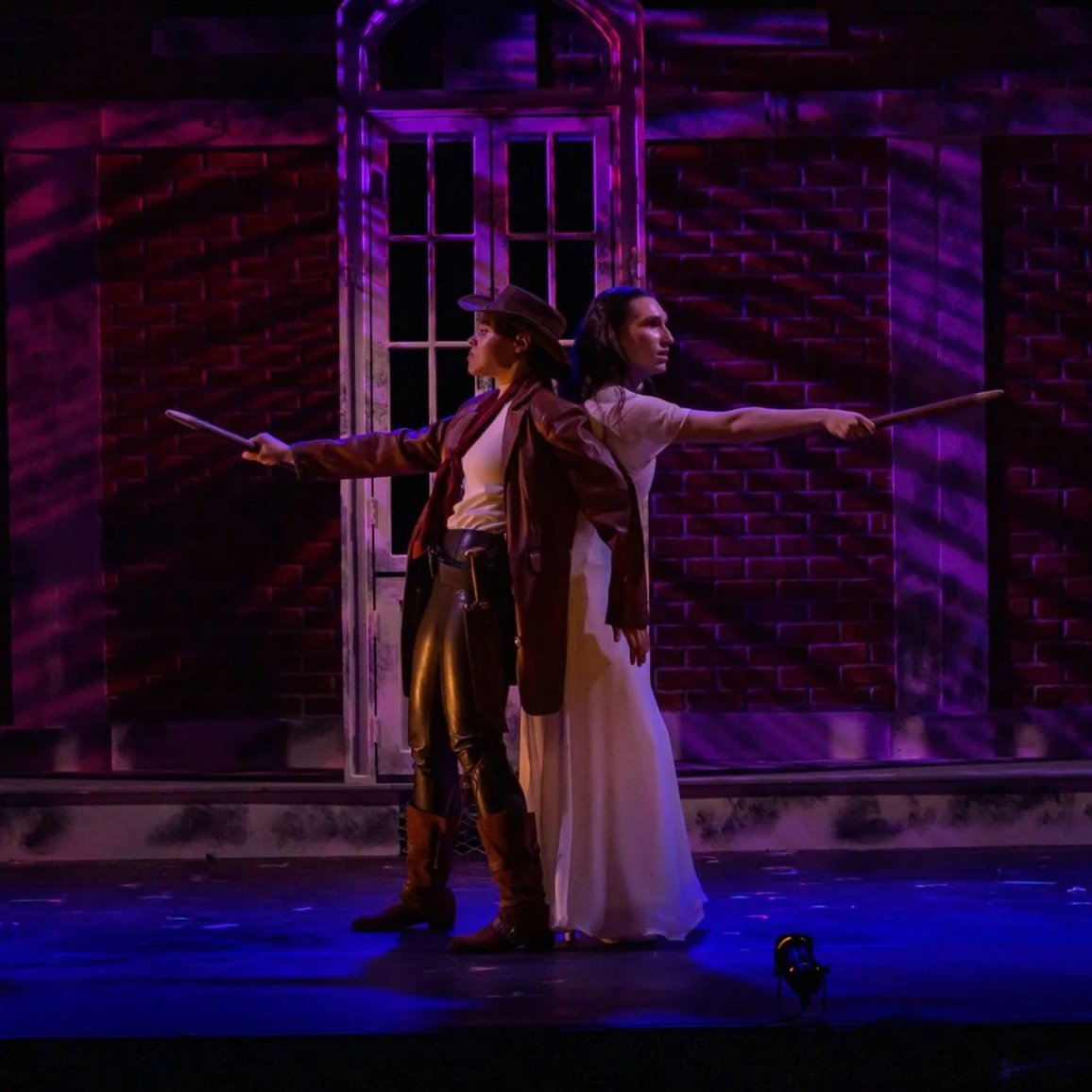 Two women stand back-to-back on stage, holding sticks, with a brick wall and a window in the background, illuminated by purple and pink stage lights.