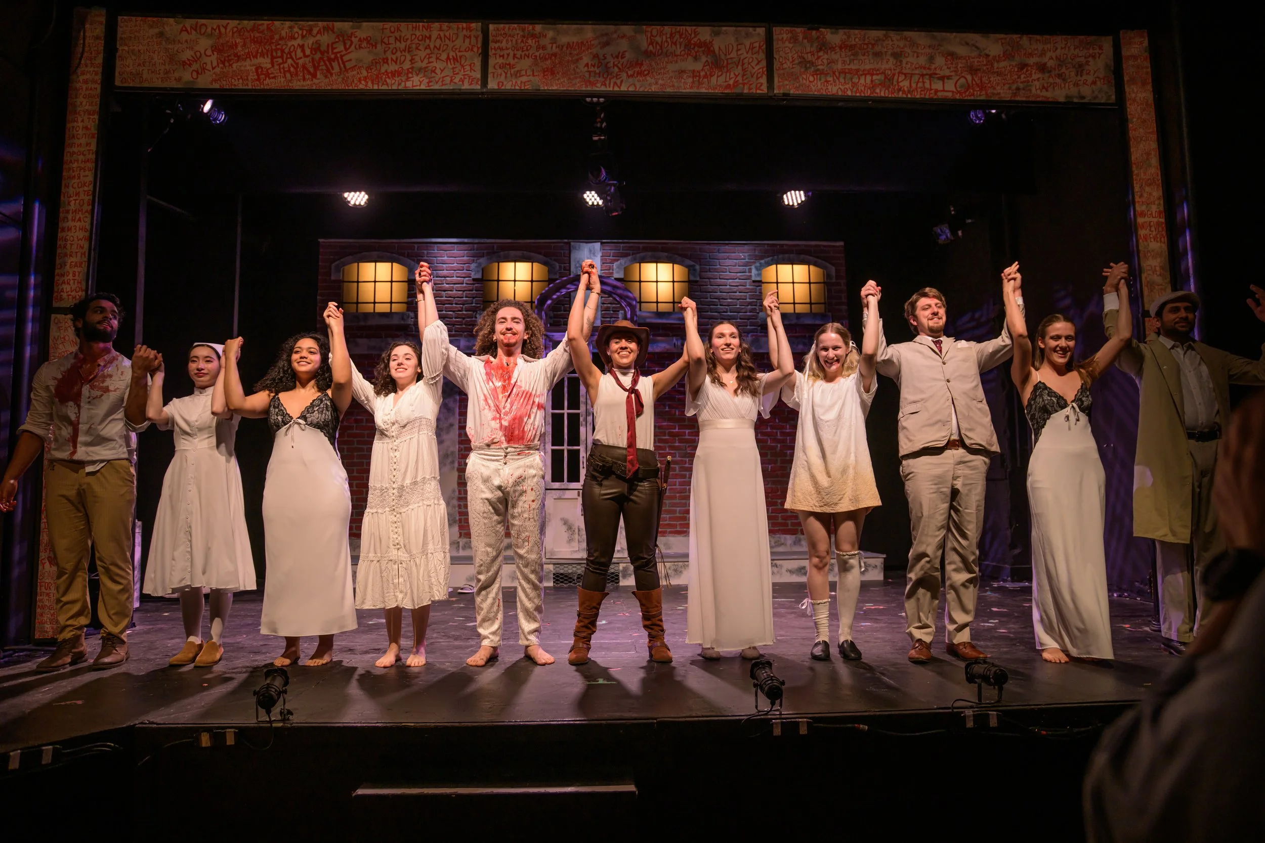 Cast of a theatrical play taking a bow on stage after a performance, holding hands and smiling. The stage has a brick wall backdrop with windows, and there is stage lighting.