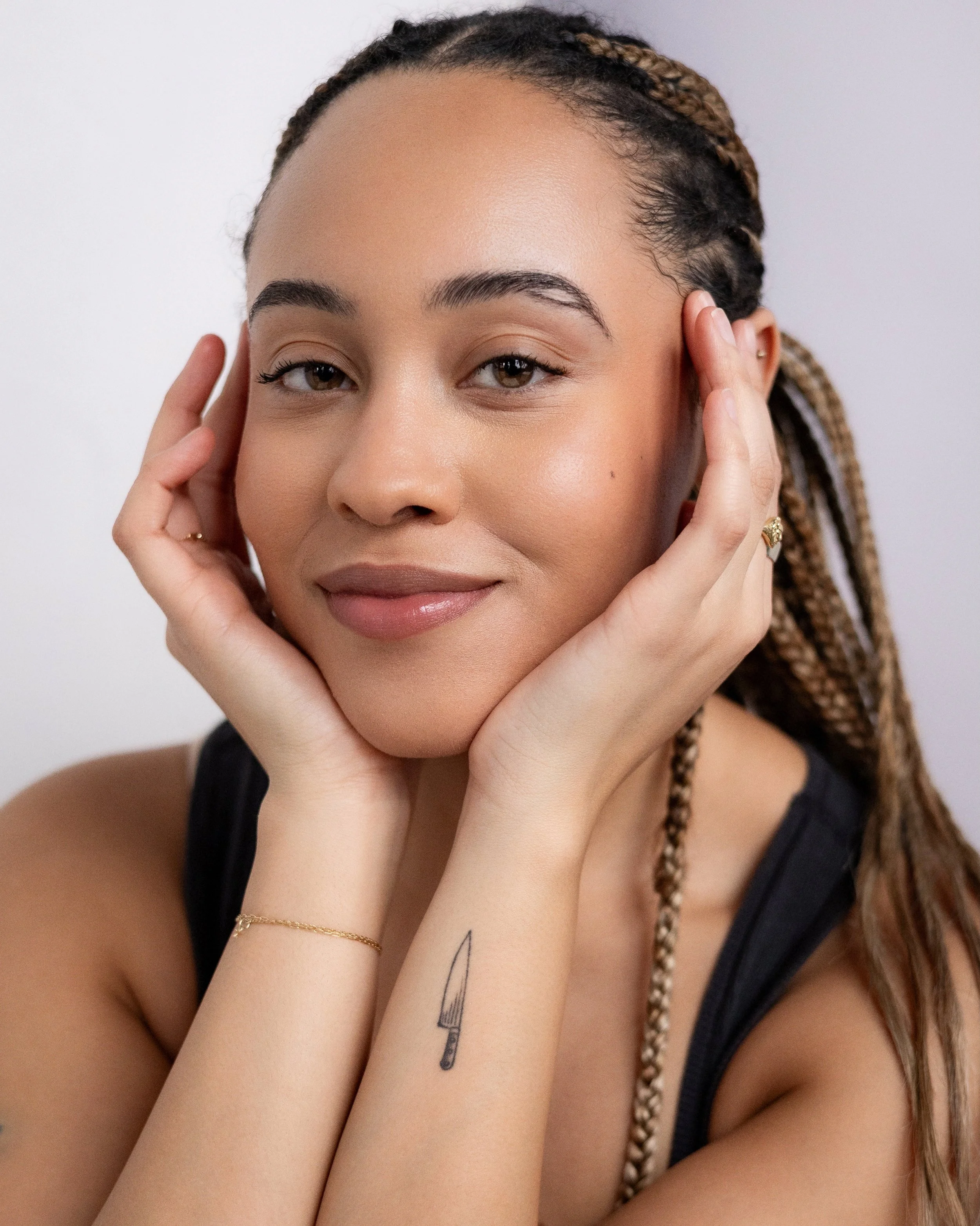 Close-up of a young woman with braided hair, smiling softly, touching her face, wearing a black sleeveless top, a thin gold bracelet, and a small tattoo of a chef's knife on her wrist.