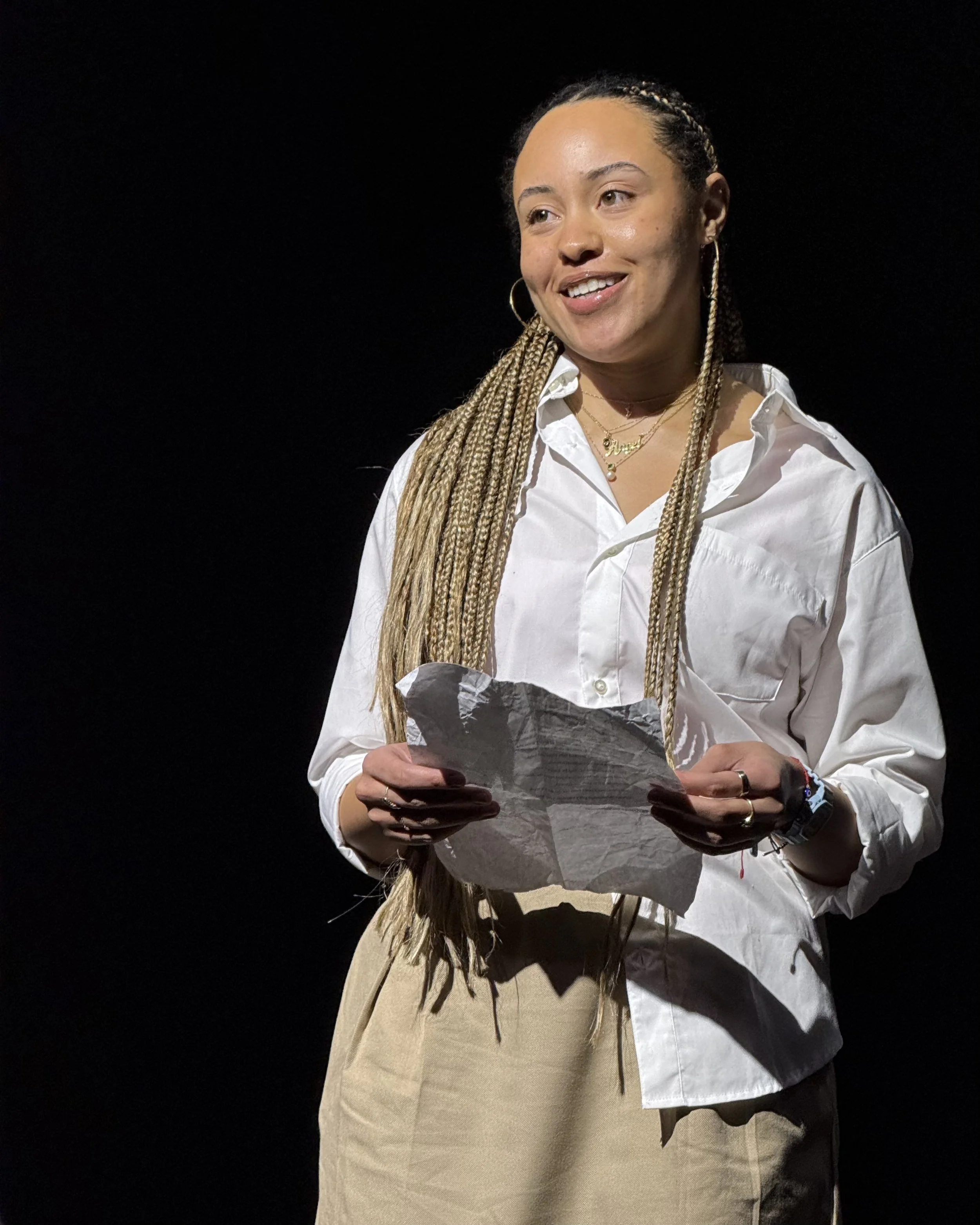 A woman with long braids holding a crumpled piece of paper, wearing a white button-up shirt and beige pants, standing against a dark background.