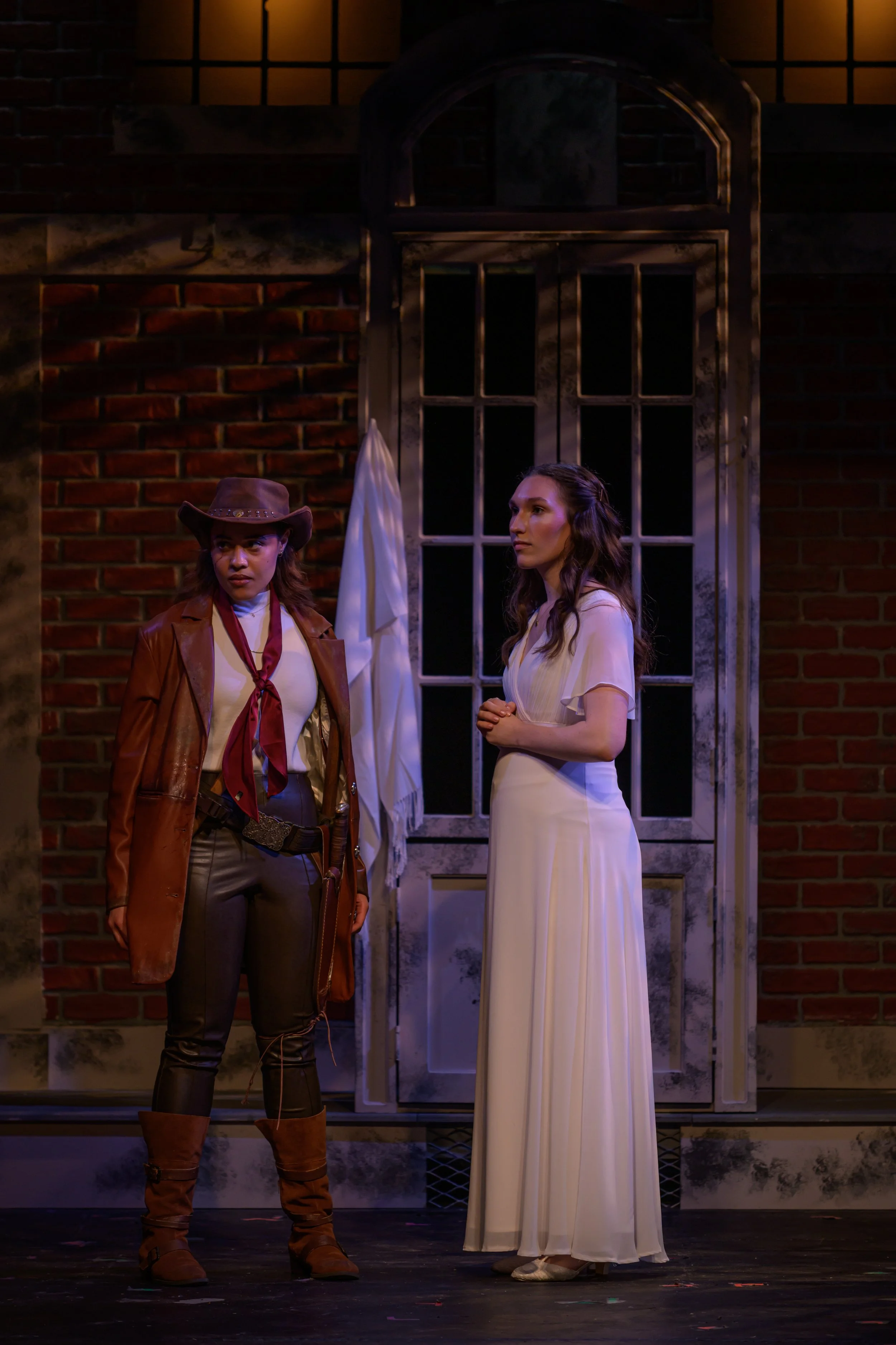 Two women on stage, one dressed as a cowgirl and the other in a white dress, in front of a brick wall and tall window, likely performing in a play.