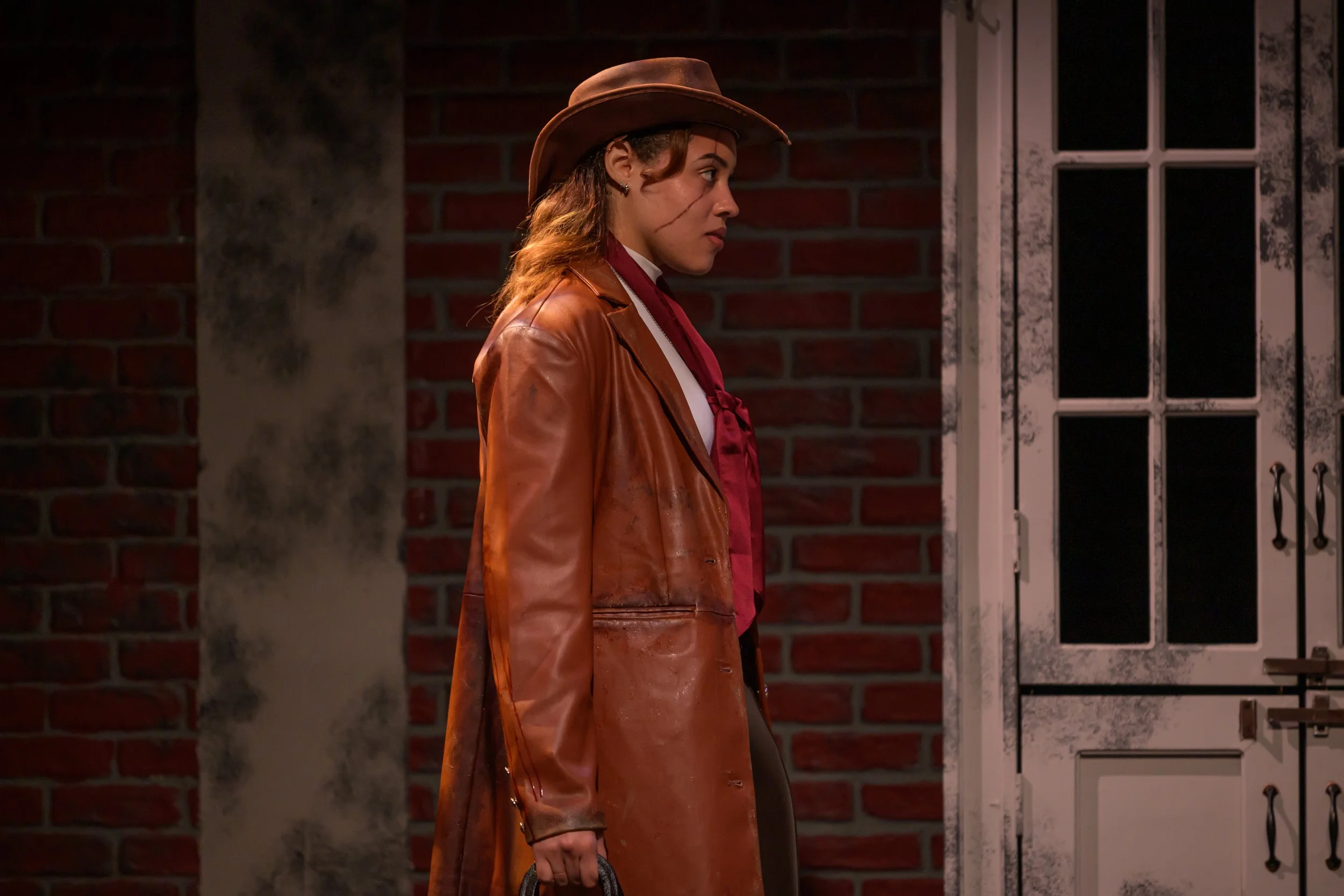 A woman dressed as a cowboy with a brown hat and leather coat standing in front of a brick wall and a door.