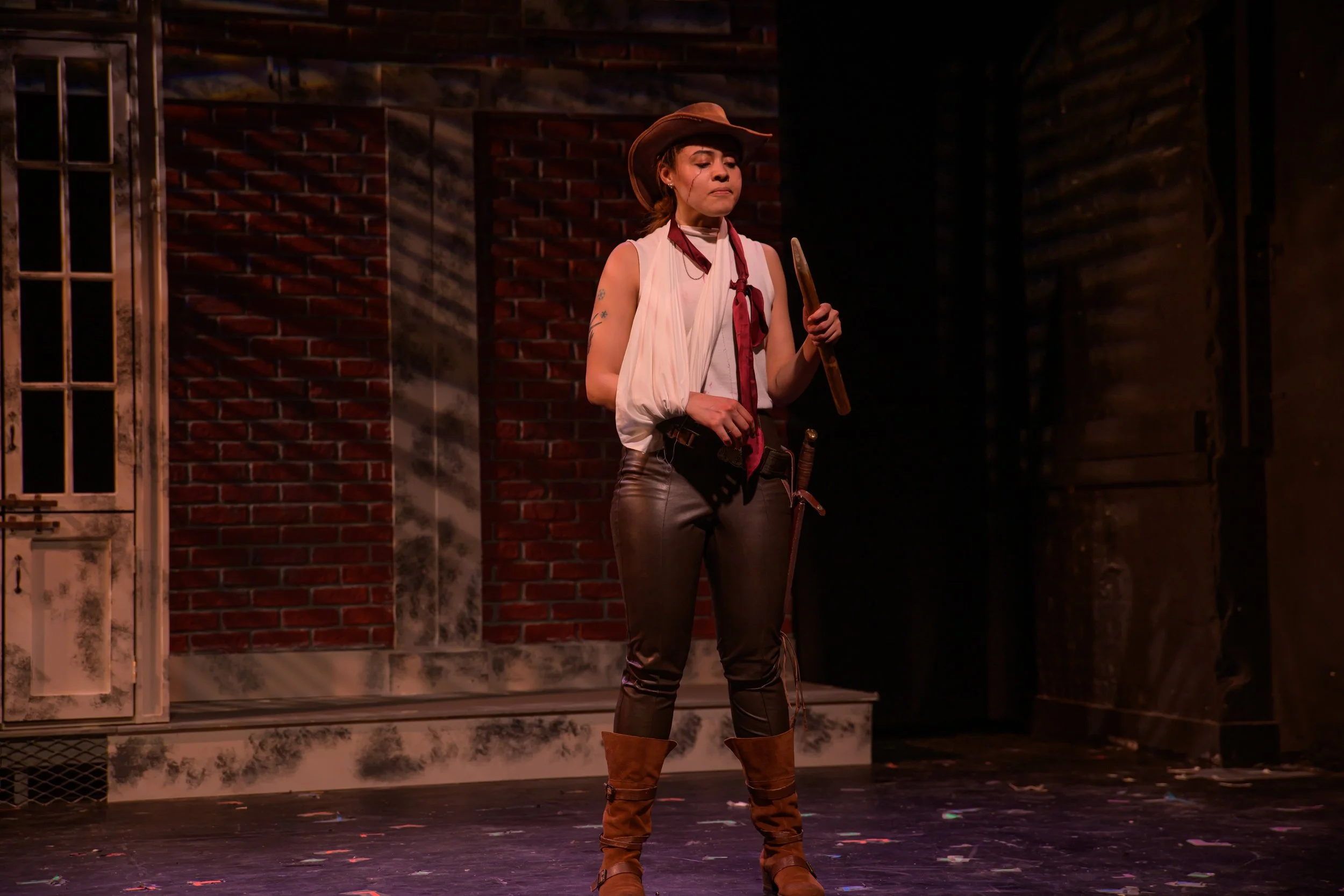 A woman dressed as a cowboy standing on a stage with a brick wall backdrop, holding a stick or weapon, wearing leather pants, a white blouse, a red scarf, a cowboy hat, and brown boots.