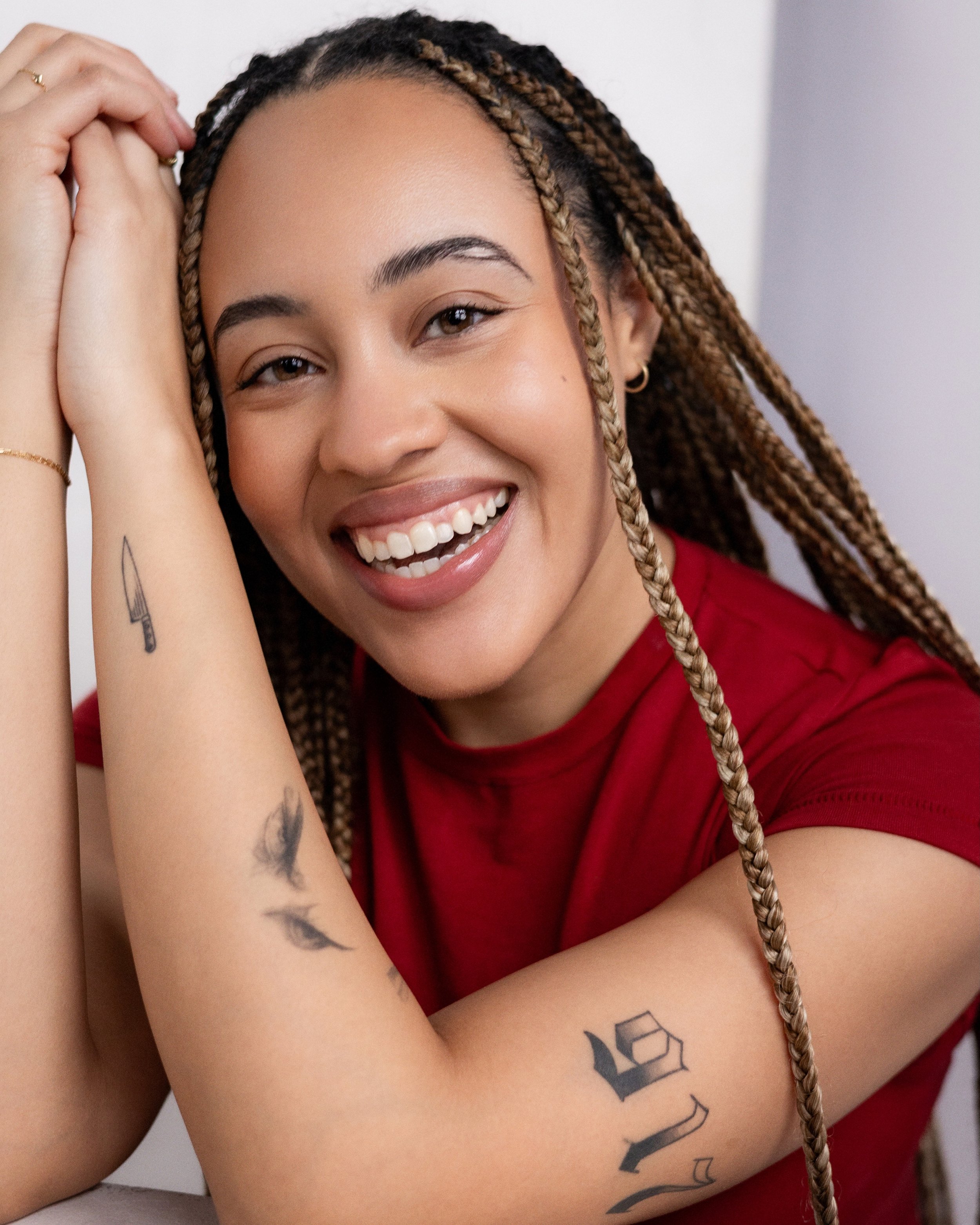A young woman smiling with braided hair, wearing a red shirt, and showing tattoos on her arm.