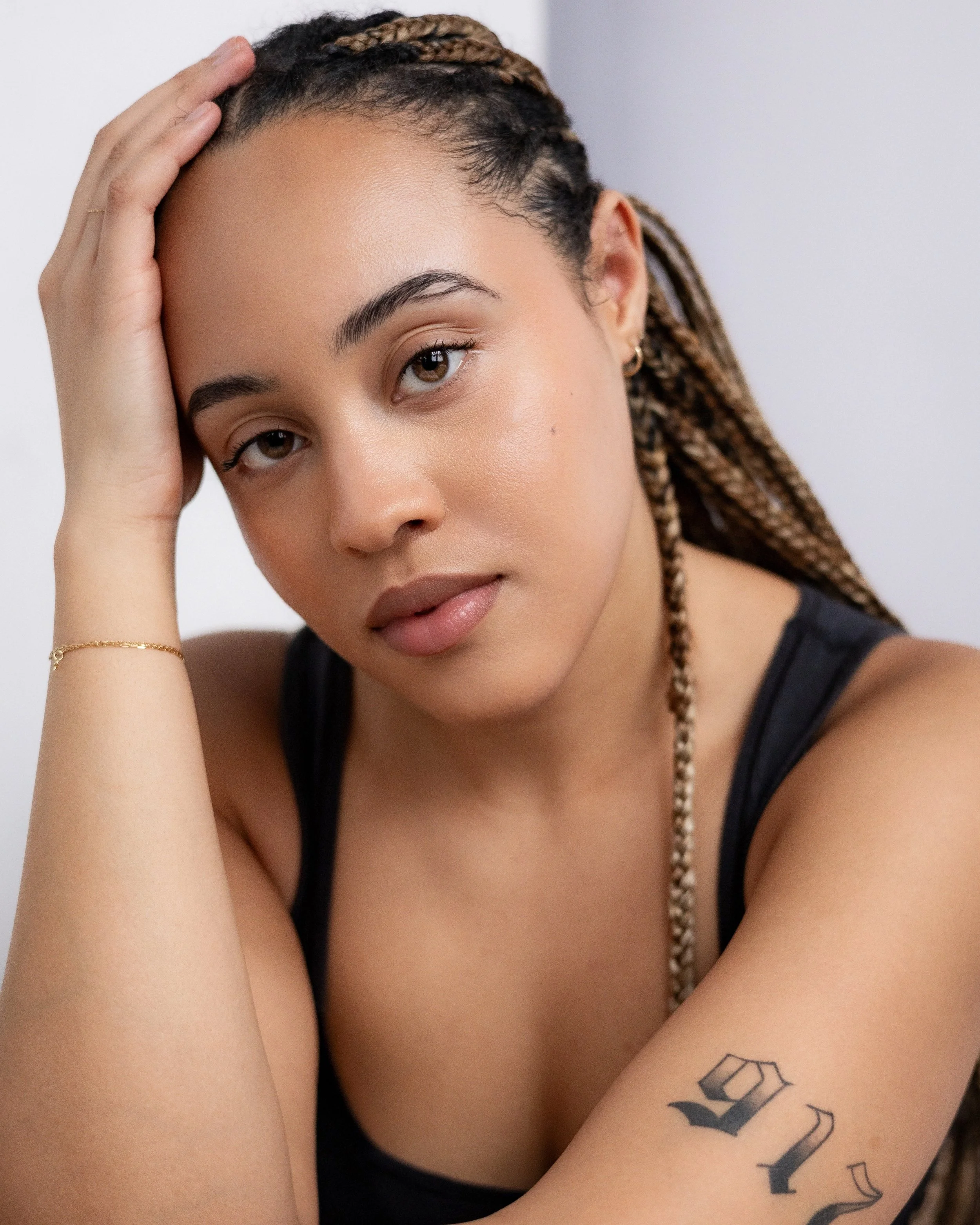 A young woman with braided hair, wearing a black tank top, resting her head on her hand, with a neutral expression and a tattoo on her arm.