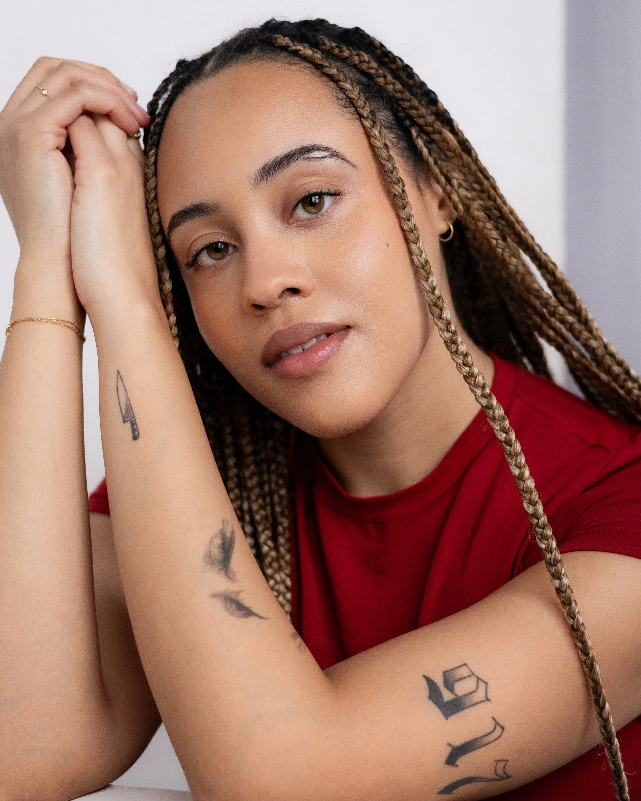 Close-up of a young woman with braided hair, wearing a red shirt, looking at the camera with a gentle expression, resting her head on her arms, showing tattoos on her left arm.