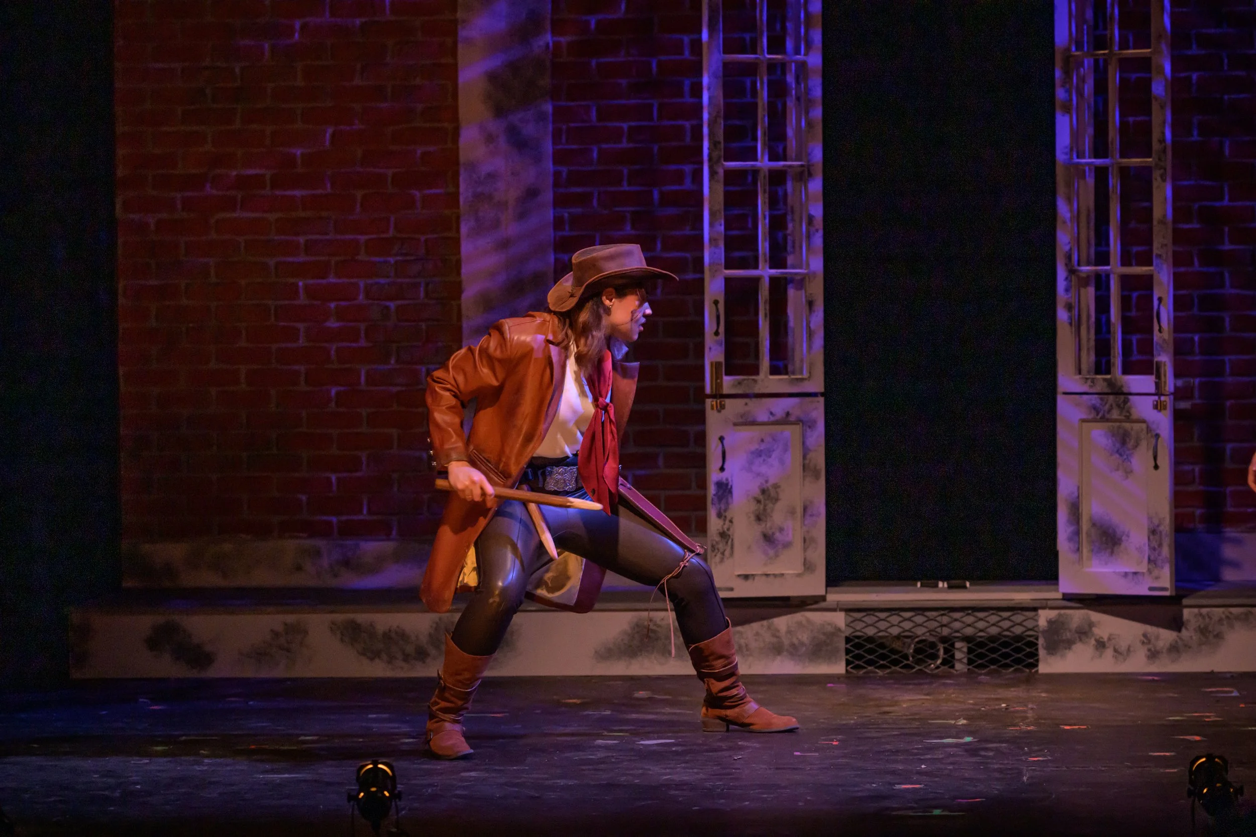 Performer dressed as a cowboy in a theatrical production, holding a hatchet, in a stance on stage with brick backdrop and open window props.