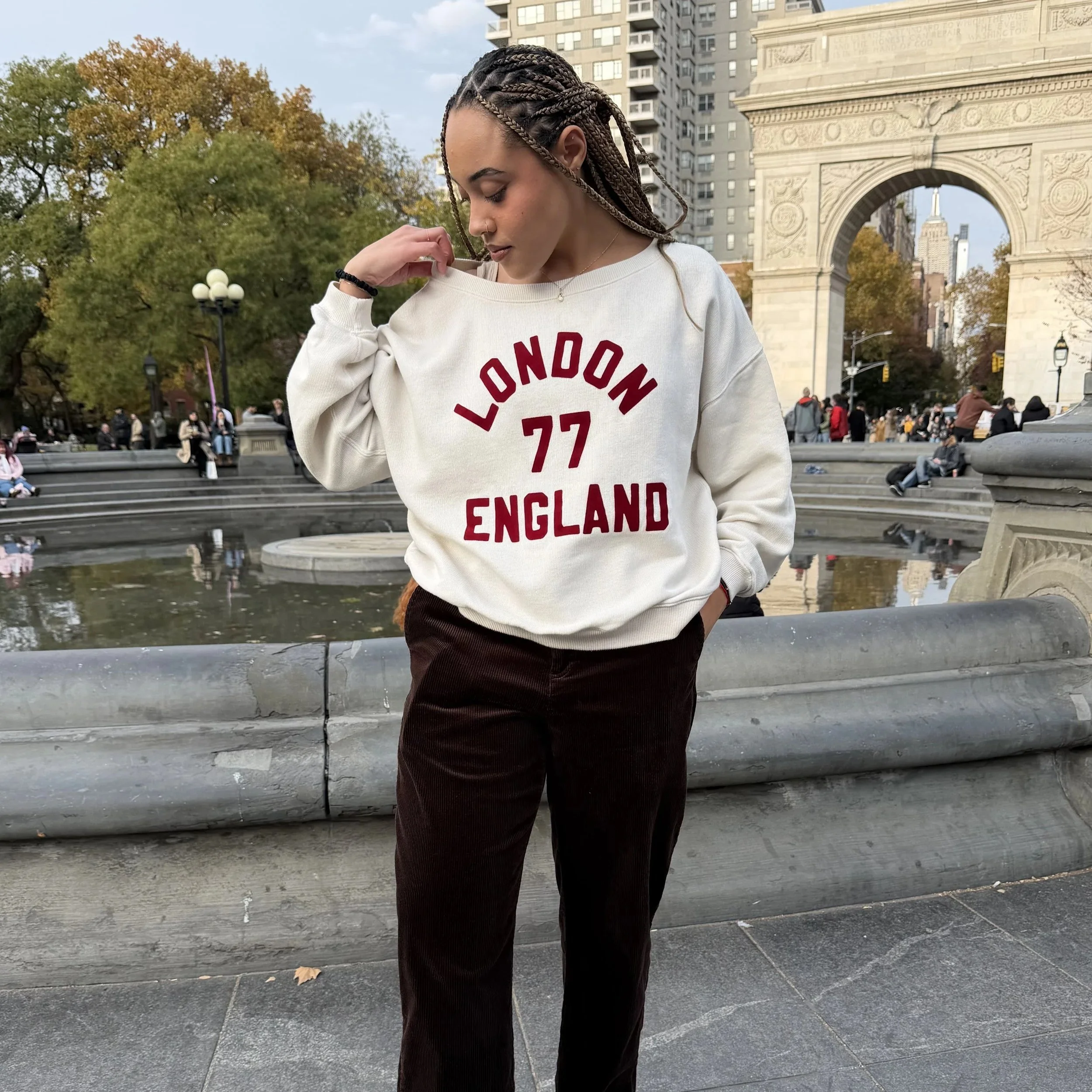 A young woman stands in front of a fountain in a city park, wearing a cream-colored sweatshirt with 'LONDON 77 ENGLAND' in red letters and dark pants, with trees and city buildings and a large arch structure in the background.