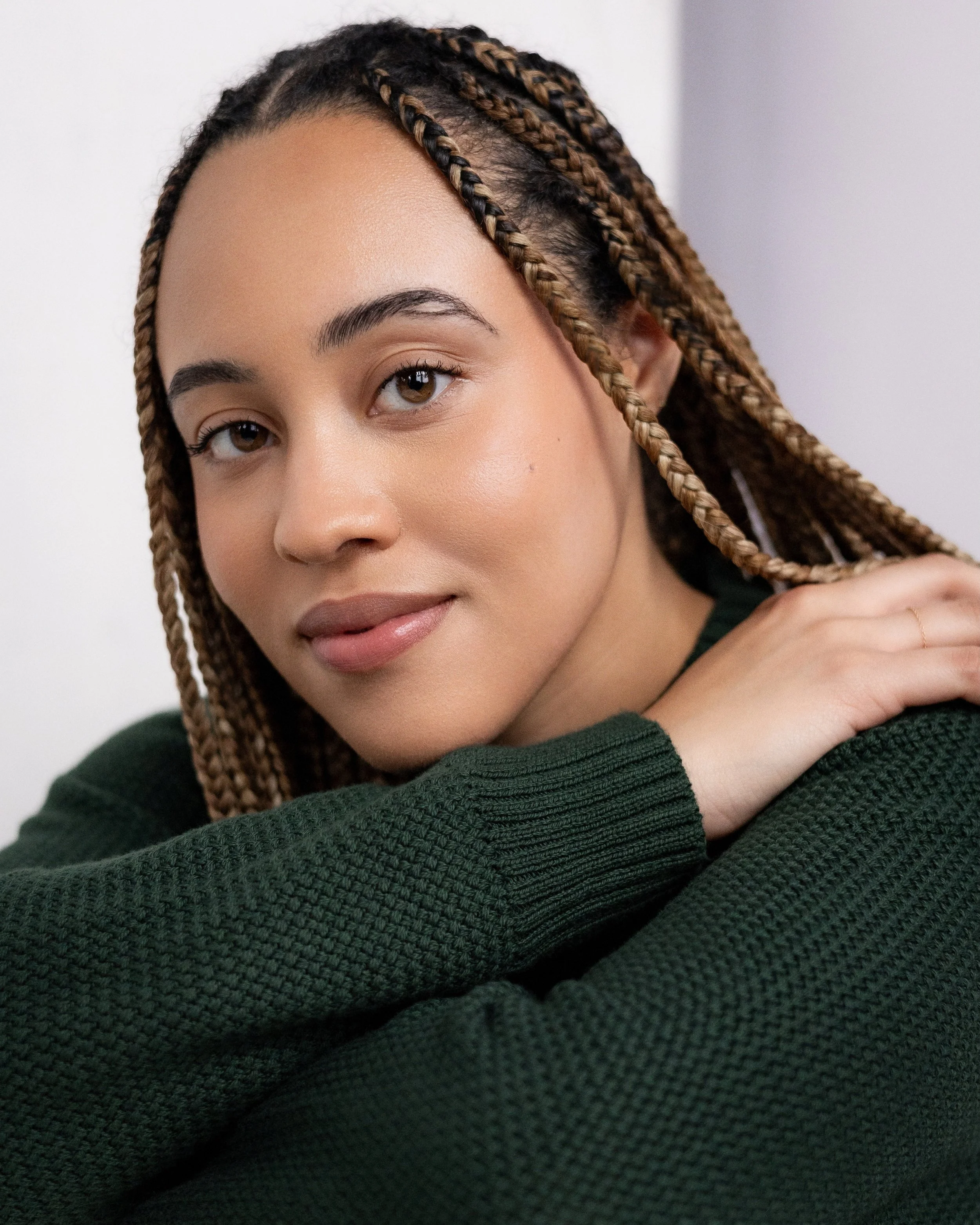 Close-up of a young woman with brown braided hair, wearing a dark green sweater, looking at the camera with a gentle smile.