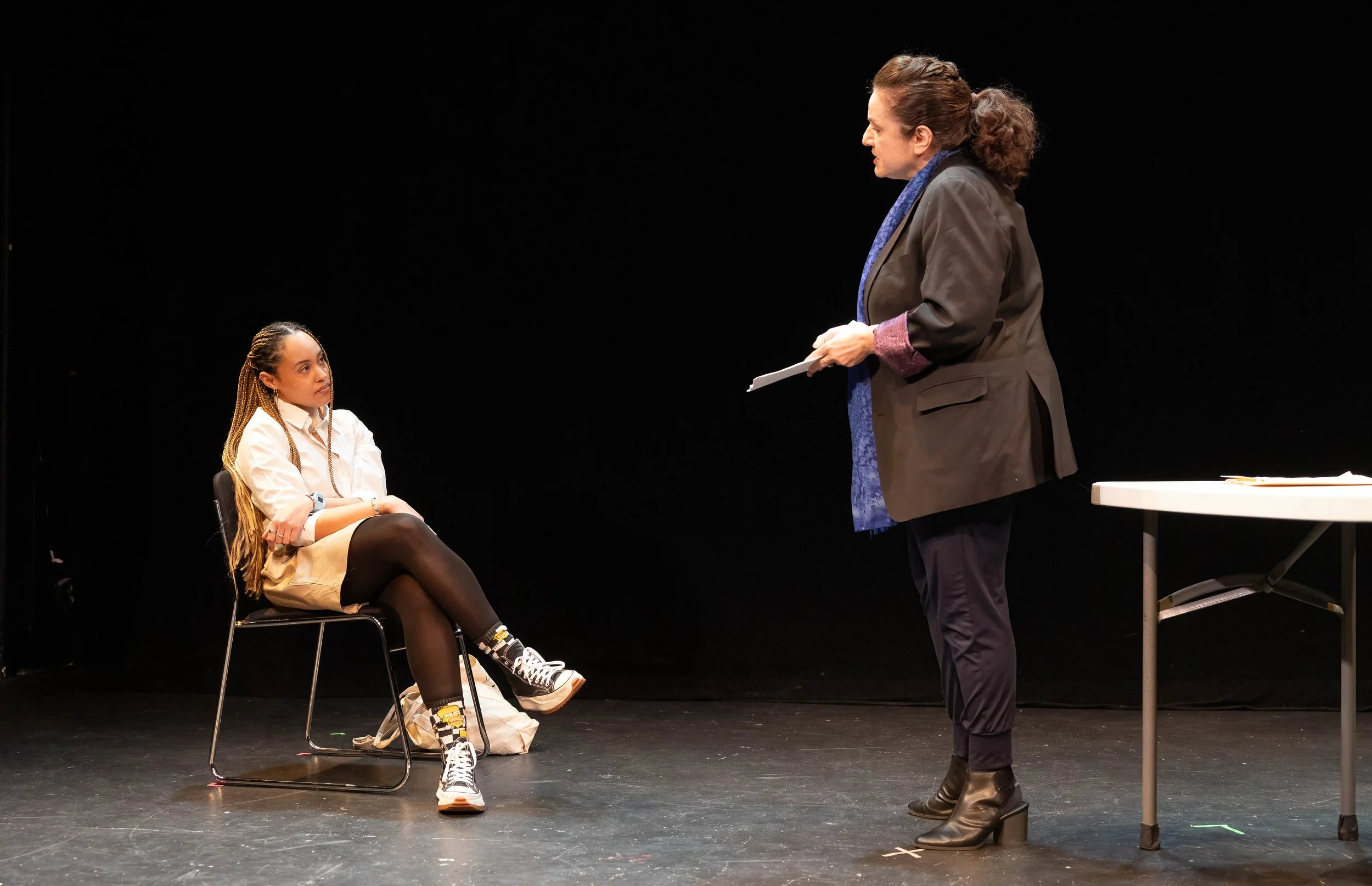 One woman sitting on a chair and another woman standing and talking to her on a stage.