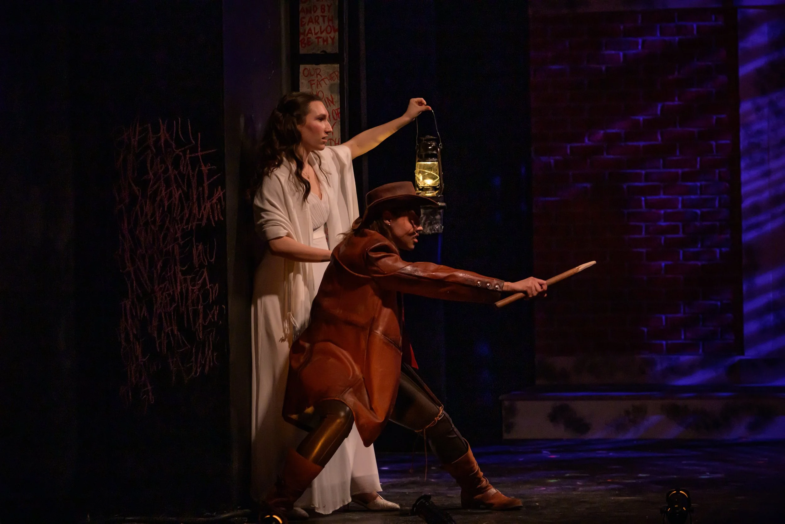 Two women are on stage in costume. One woman, standing in a white dress, holds a lantern aloft. The other woman, dressed in a brown leather jacket, hat, and boots, is kneeling with a stick pointed forward. The stage background includes a brick wall a