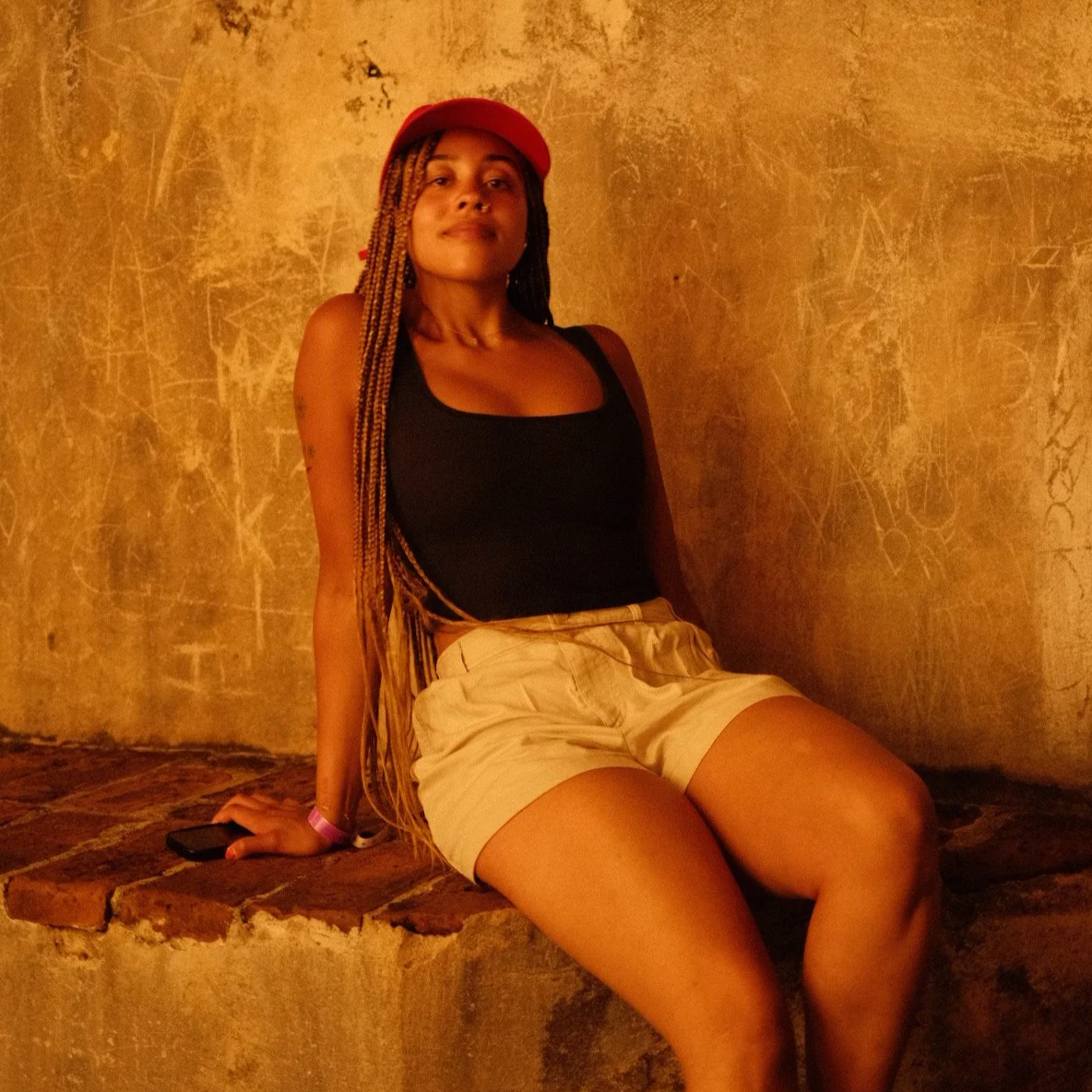A woman with braided hair and a red hat sitting on a brick ledge against a textured wall.