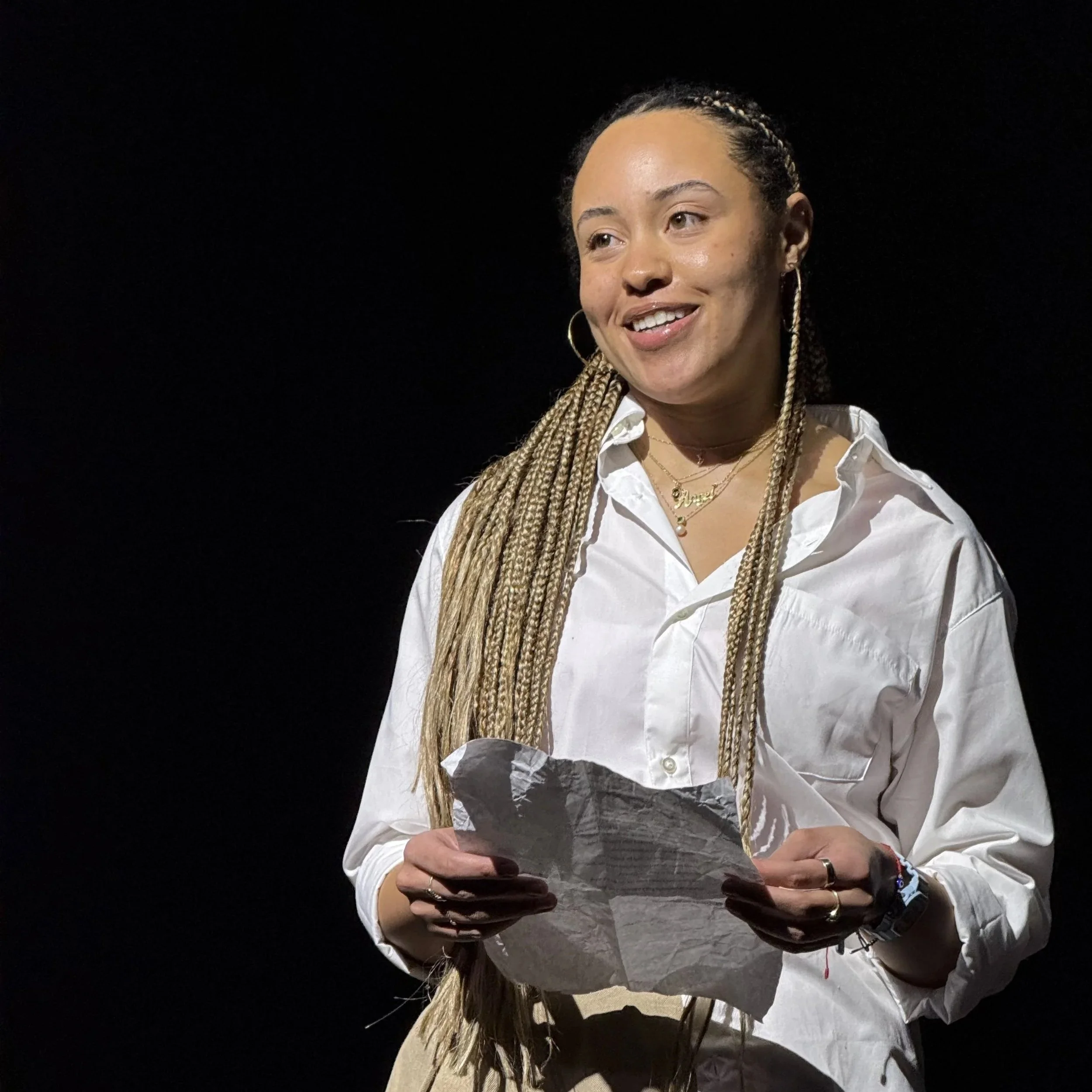 Young woman with braided hair, wearing a white shirt, holding crumpled paper, smiling, against a black background.