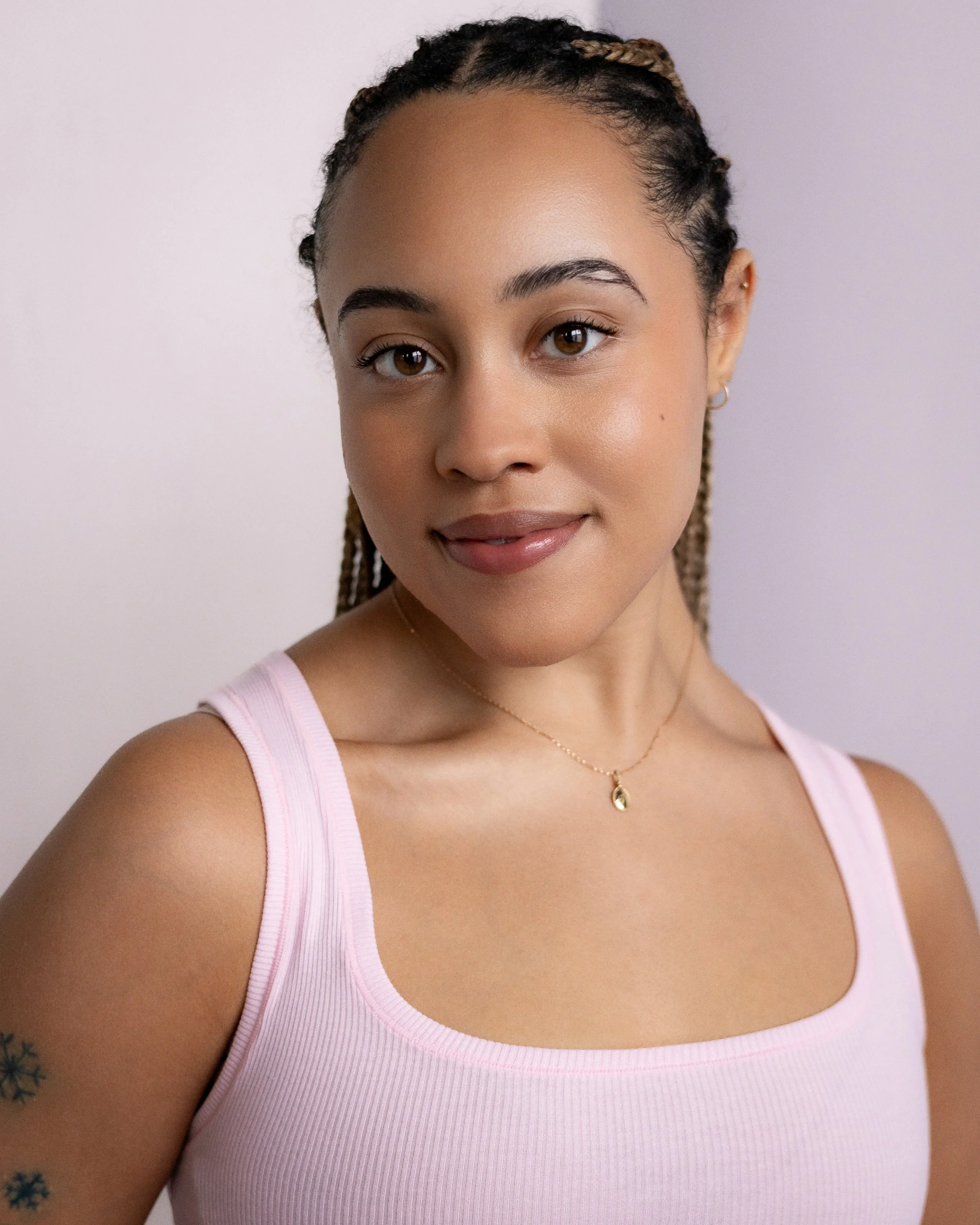 Portrait of a young woman with braided hair wearing a pink tank top and a delicate gold necklace, smiling softly against a plain background.