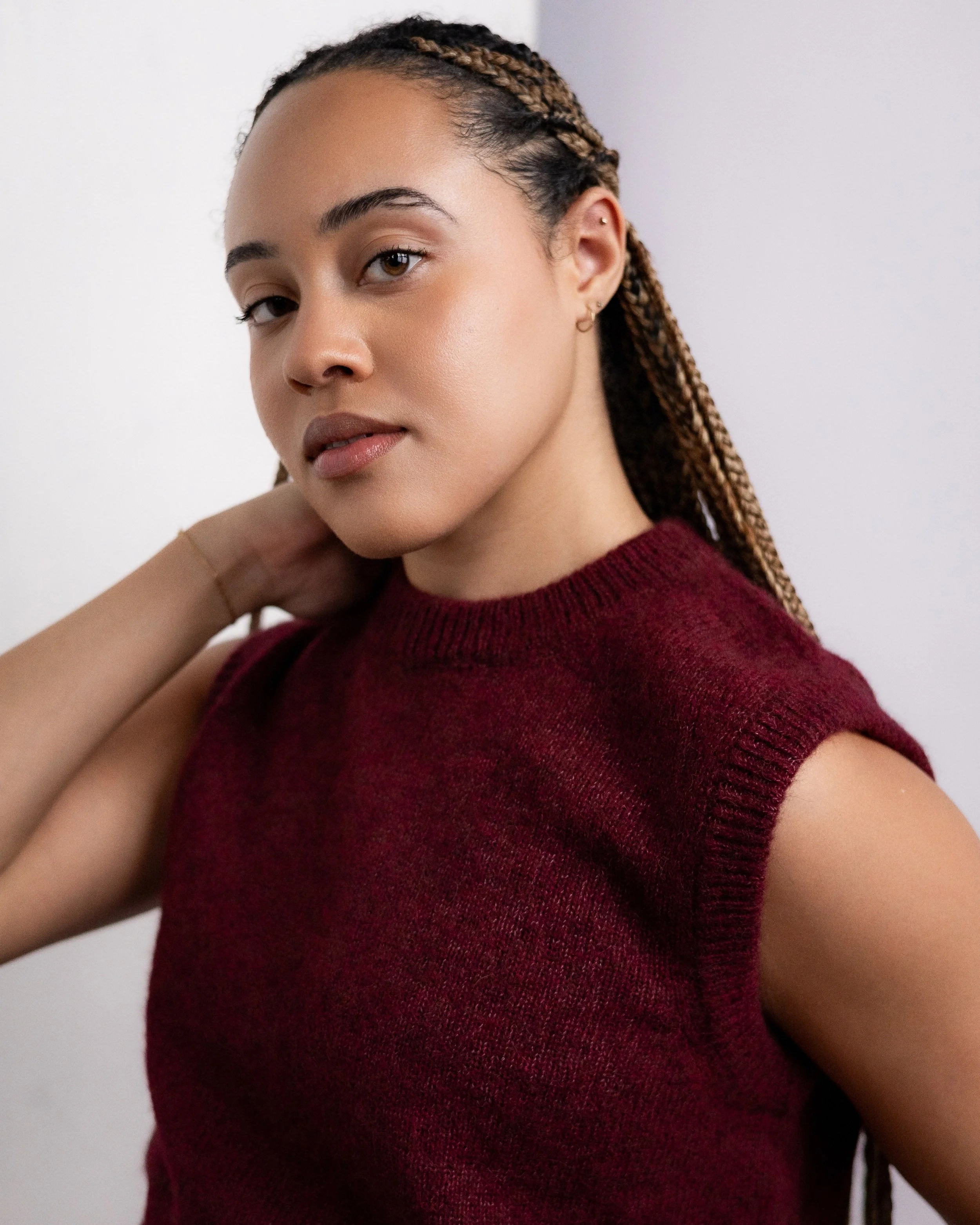 Portrait of a woman with braided hair wearing a sleeveless maroon sweater, standing against a plain white background.
