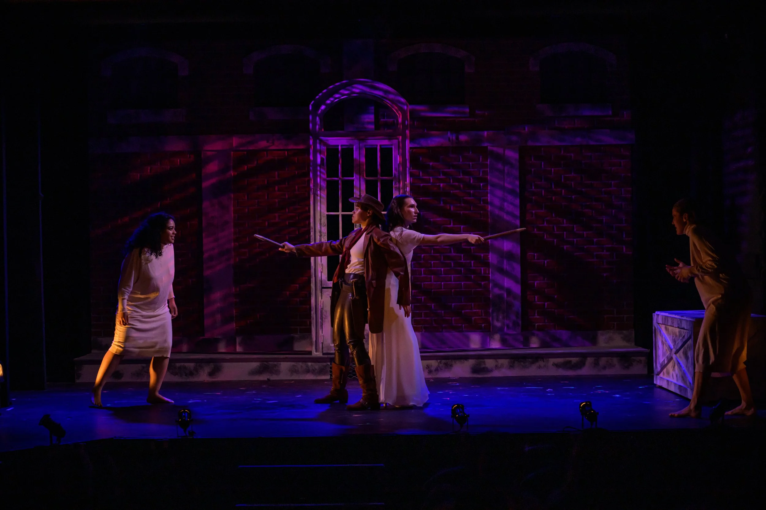 Actors performing a theater scene on stage with a brick wall background and a window, illuminated by purple stage lighting.