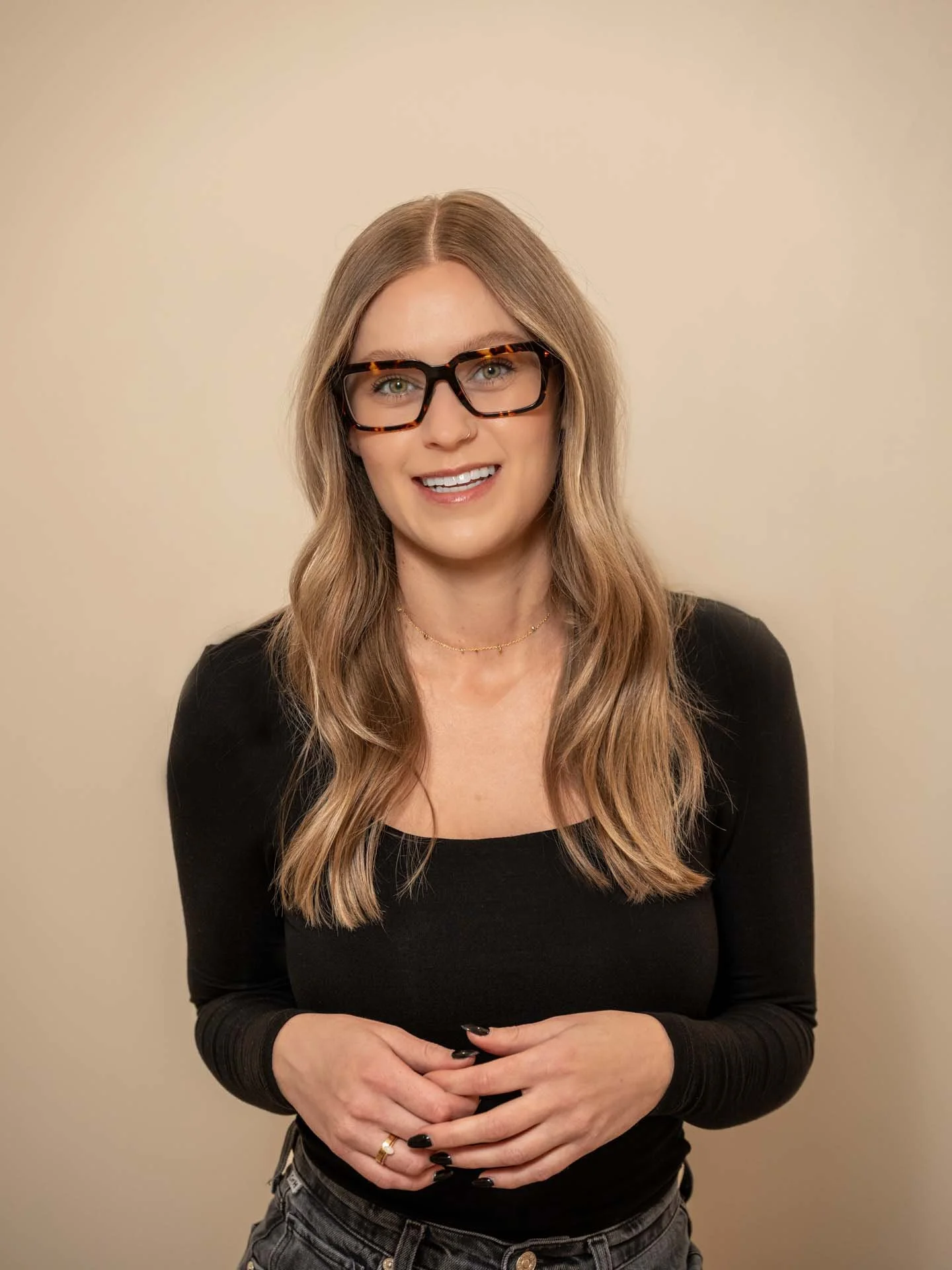A woman with long wavy blonde hair, wearing a black top and stylish glasses with a tortoiseshell pattern, smiling and standing against a plain beige background.